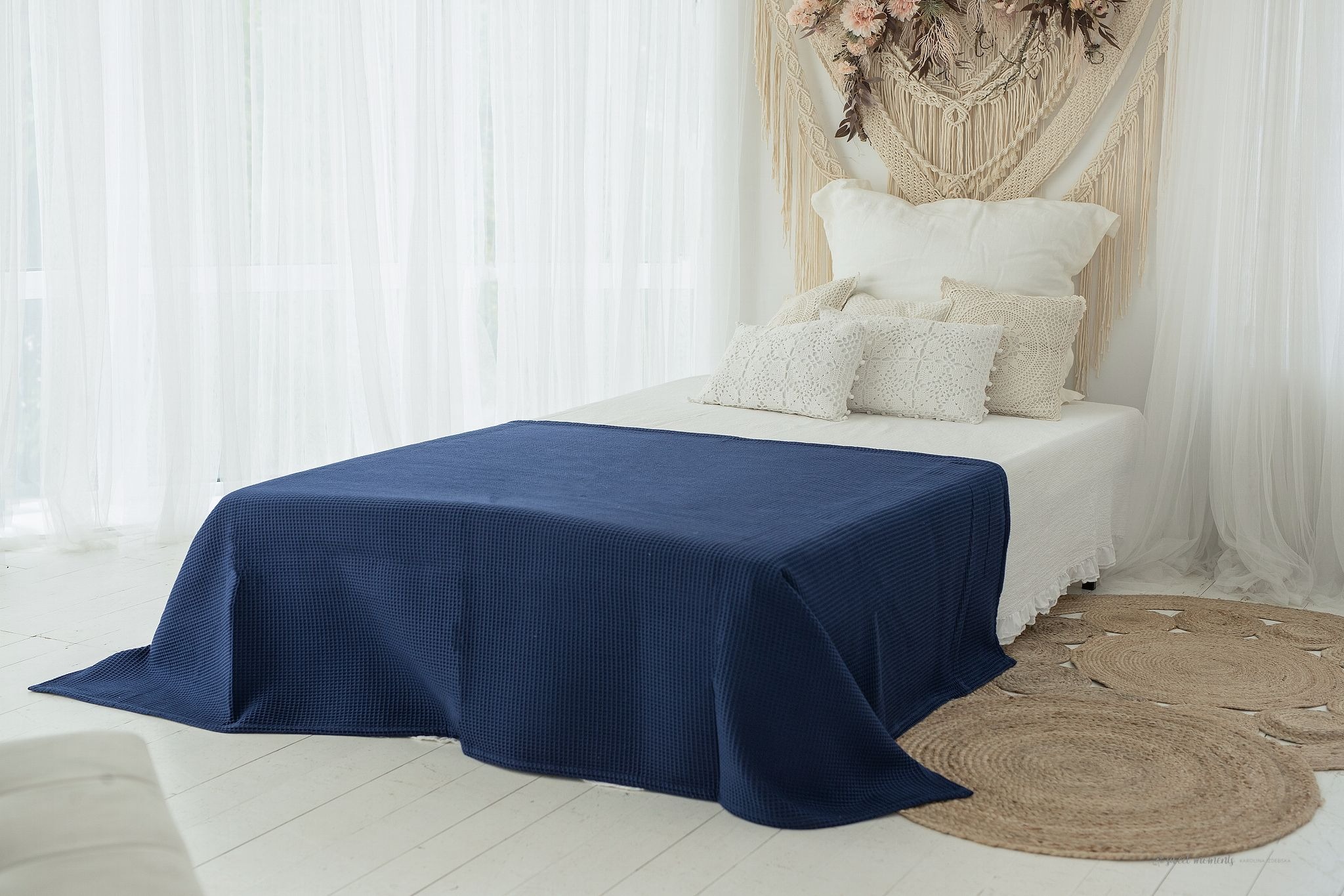 Niebieska narzuta wafelkowa na łóżku z białymi poduszkami dekoracyjnymi, jutowe dywaniki i makrama z kwiatami w tle, przy oknie z białymi zasłonami.