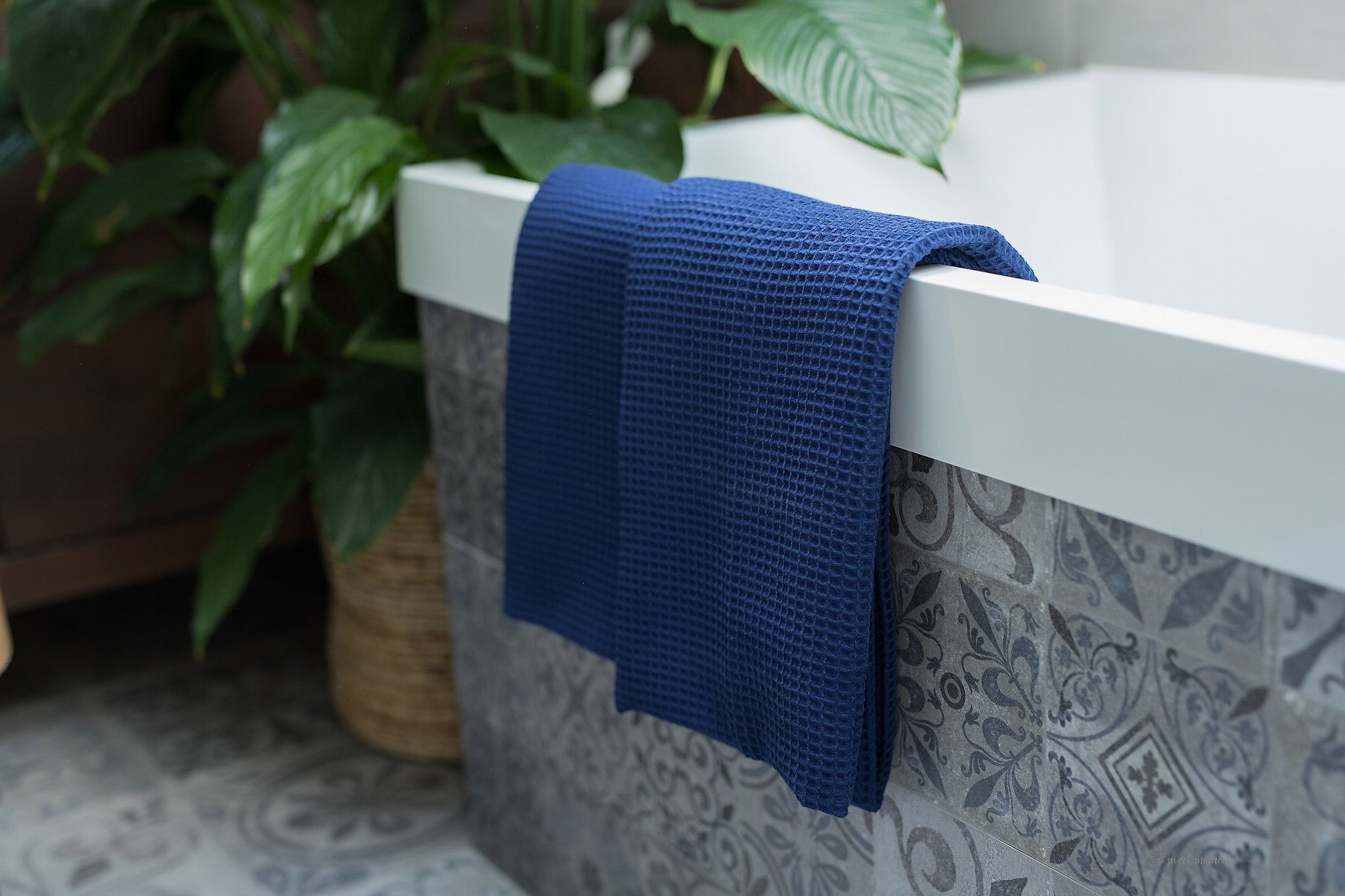 Niebieski ręcznik z tkaniny wafelkowej zawieszony na białej krawędzi wanny z szarymi, wzorzystymi płytkami, z rośliną doniczkową w tle.