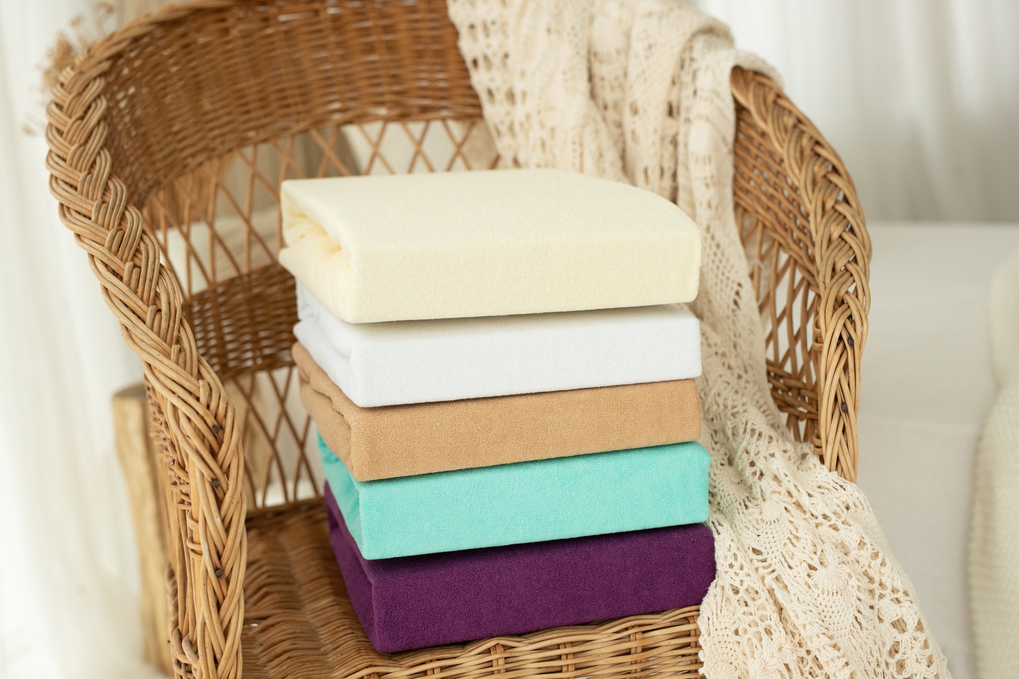Stos prześcieradeł z gumką w kolorach: kremowym, białym, beżowym, turkusowym i fioletowym, ułożony na wiklinowym fotelu z ażurowym pledem w tle.