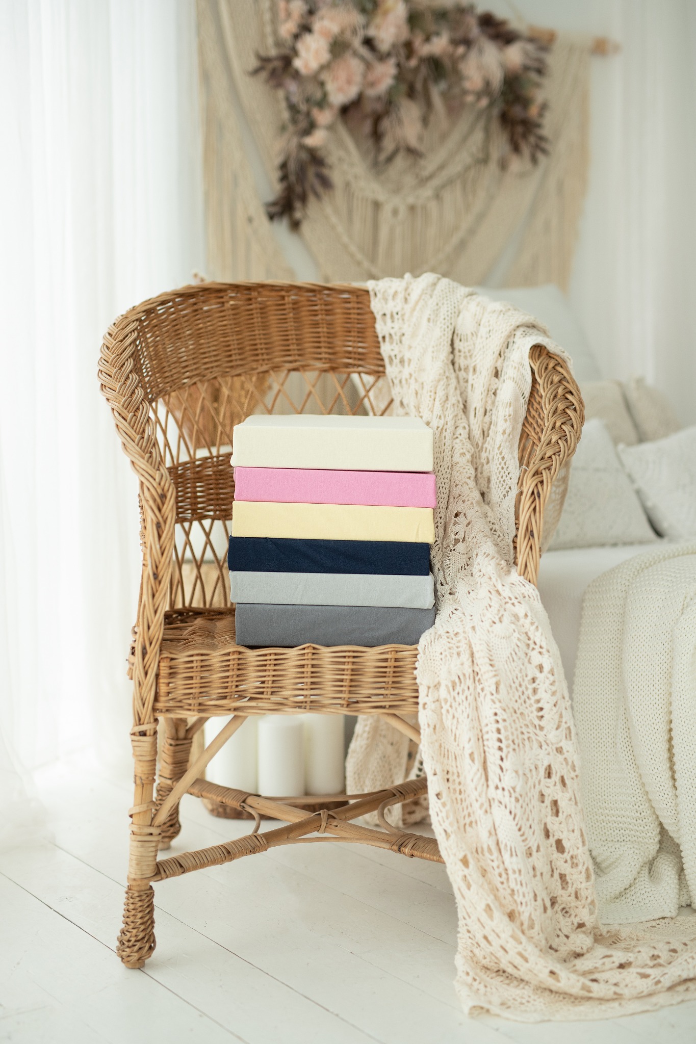 Stos kolorowych prześcieradeł złożonych na wiklinowym krześle, z dekoracyjnym pledem i makramą w tle, w jasnym wnętrzu.