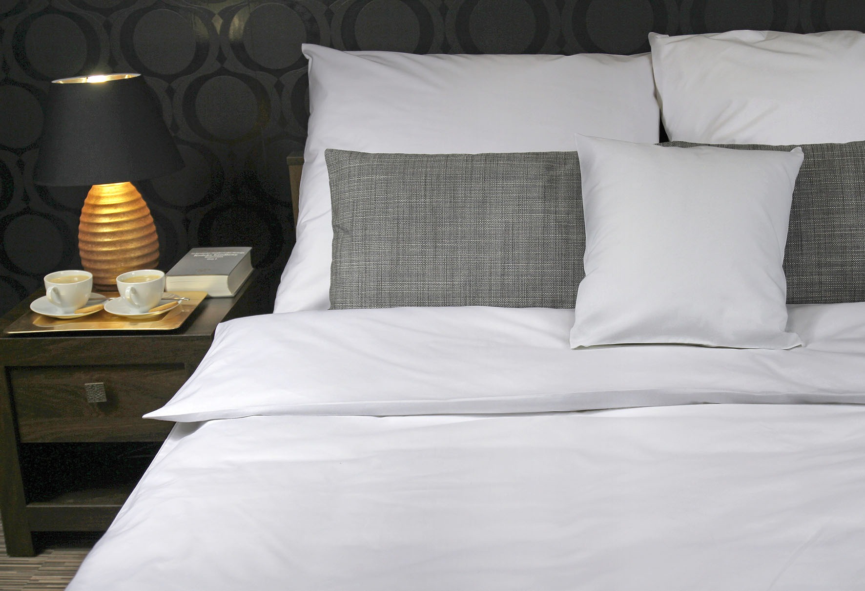 Biała pościel hotelowa z szarymi poduszkami dekoracyjnymi na łóżku, stolik nocny z lampką, filiżanki z kawą i książka.