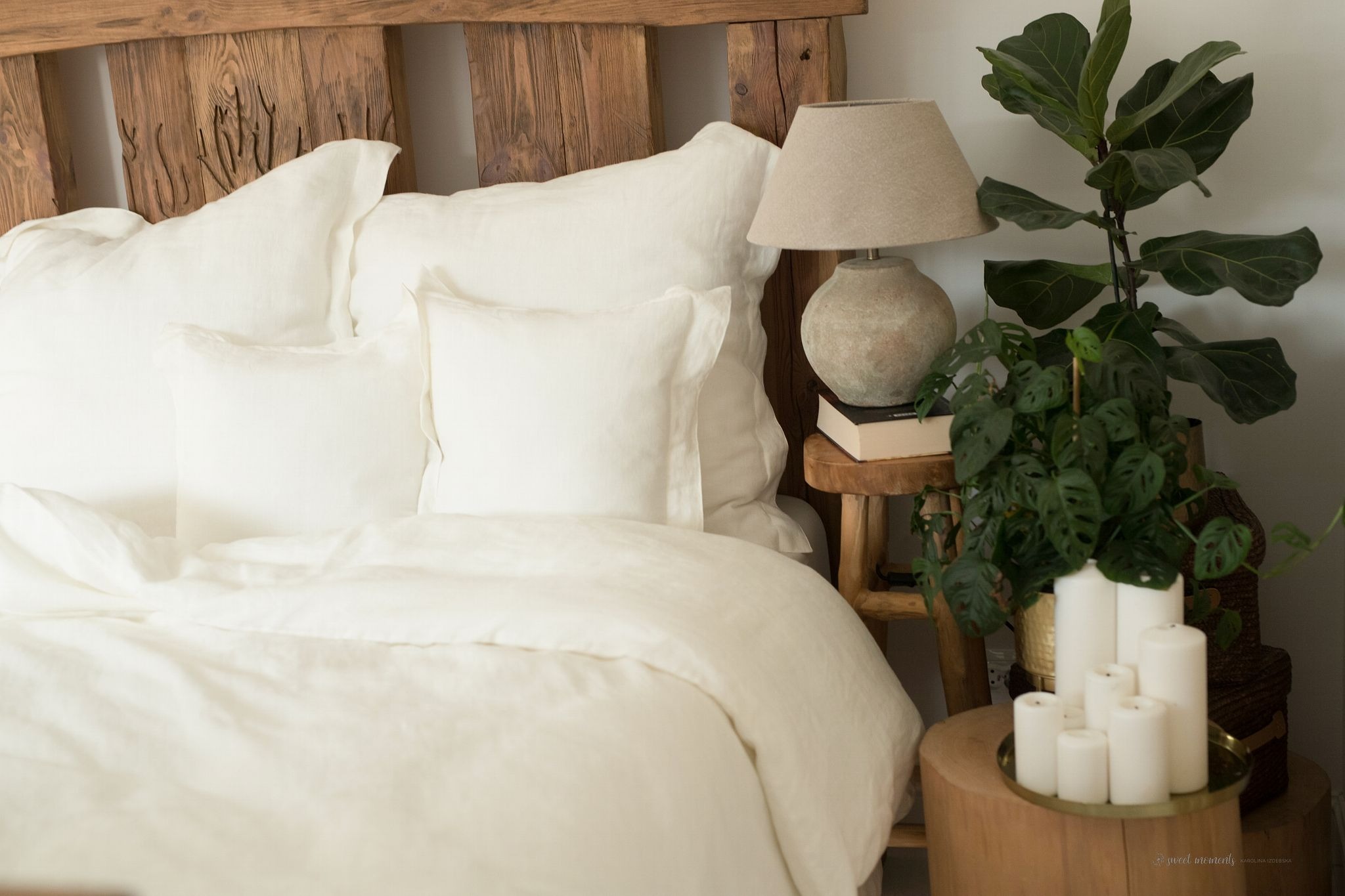 Biała, lniana pościel na łóżku z drewnianym zagłówkiem, obok stolik z lampką i roślinami doniczkowymi, świece na tacy.