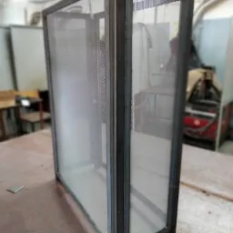 Spawana metalowa konstrukcja z siatką, widoczne spawy narożne, w tle warsztat z maszyną spawalniczą.