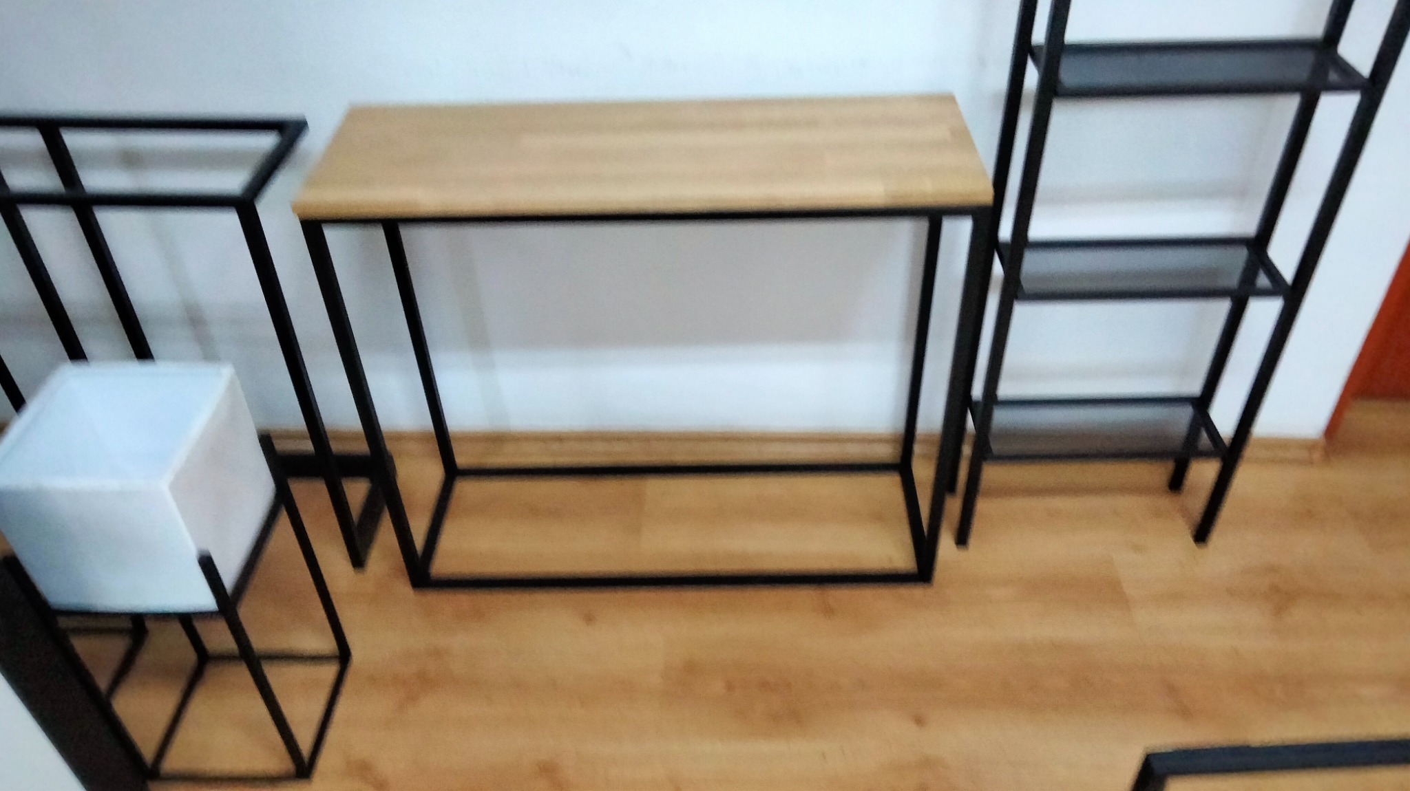 Metalowe konstrukcje meblowe: stolik z drewnianym blatem, regał i stojak na donicę, z czarnych profili, na drewnianej podłodze.
