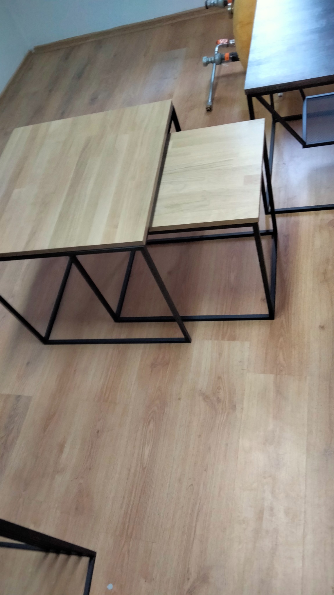 Zestaw dwóch stolików kawowych z drewnianymi blatami i czarnymi, geometrycznymi podstawami metalowymi, spawane narożniki, na panelach podłogowych.