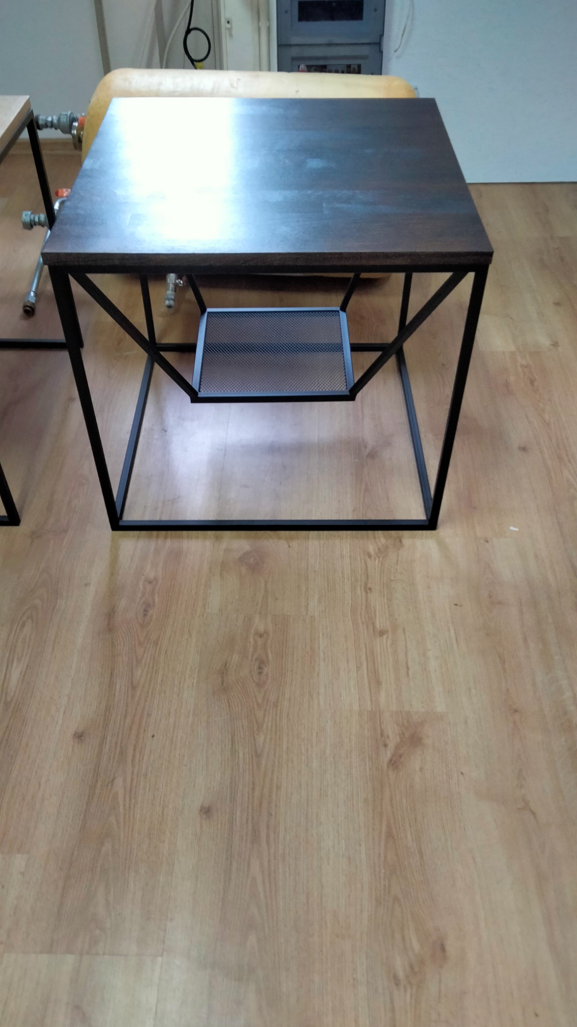 Stolik kawowy z ciemnym drewnianym blatem i czarną metalową konstrukcją, z dodatkową półką z siatki metalowej, na tle jasnej podłogi imitującej drewno.