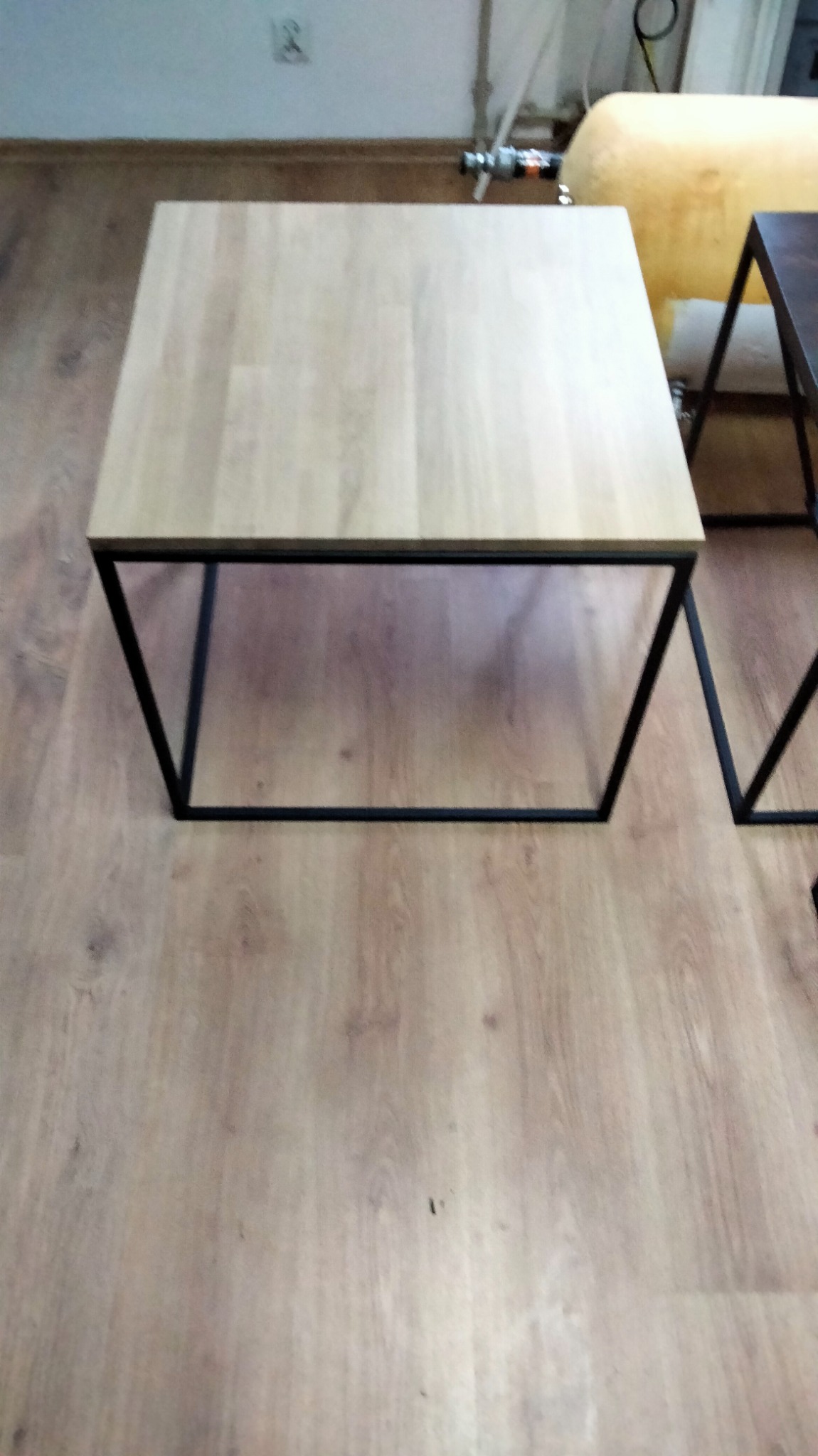 Kwadratowy stolik kawowy z jasnym blatem i czarną, spawaną podstawą z metalu, stojący na podłodze imitującej drewno.