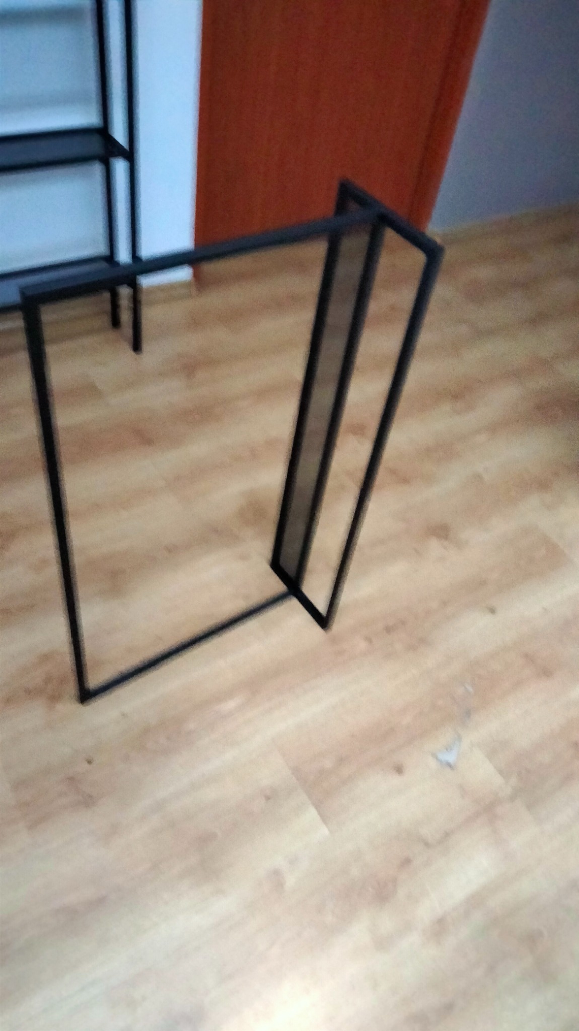 Częściowo zmontowany, czarny metalowy stelaż meblowy na drewnianej podłodze, widoczny fragment regału w tle.