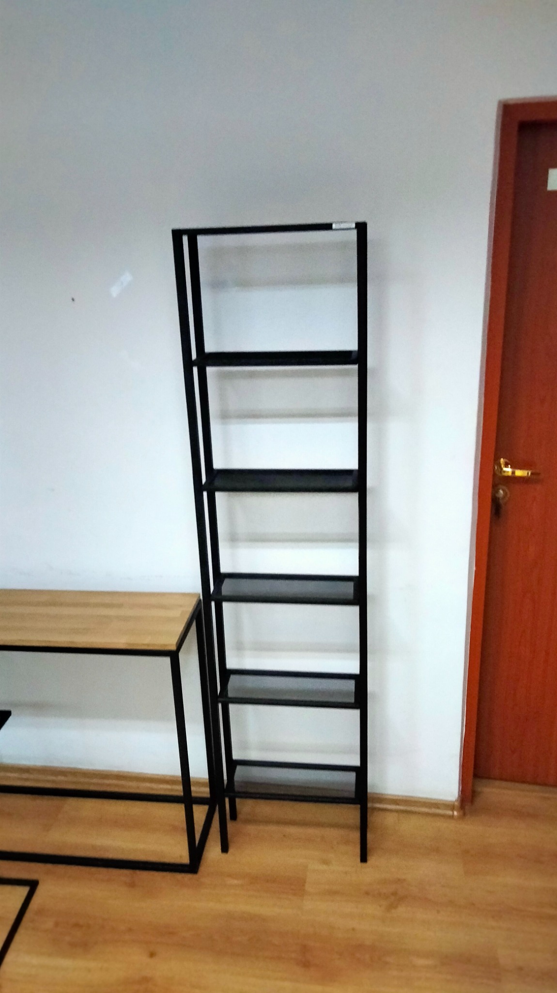 Metalowa półka regałowa z czarną ramą i drewnianymi półkami, stojąca obok drewnianego biurka z metalową ramą, na tle białej ściany i brązowych drzwi.