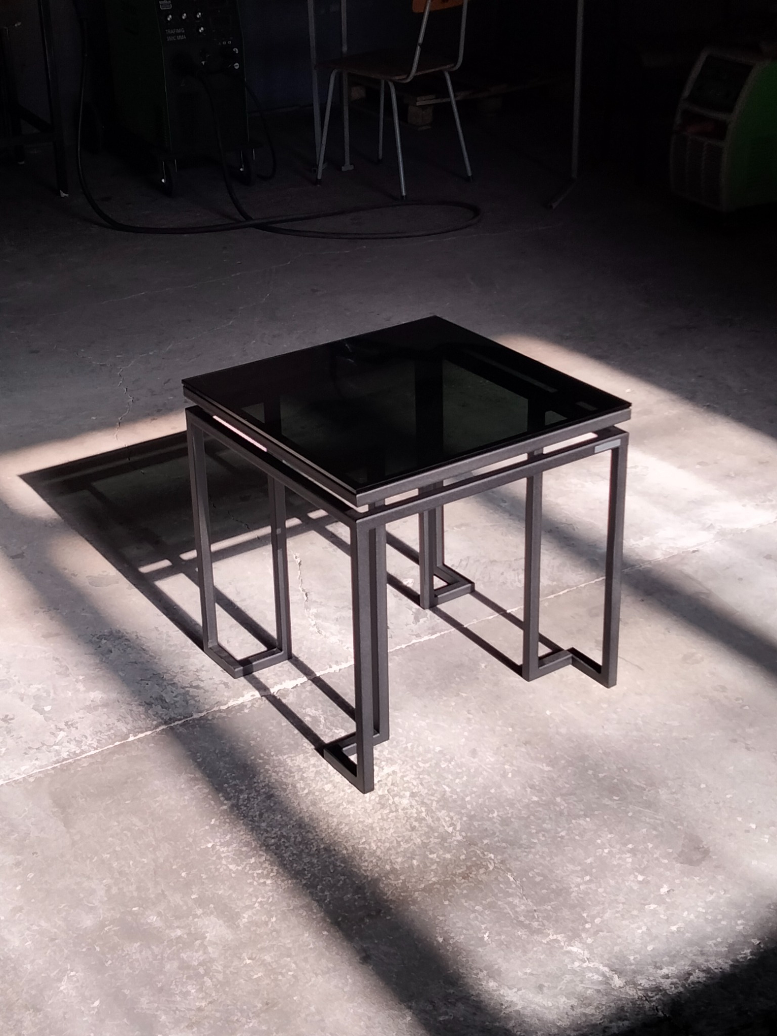 Kwadratowy stolik kawowy z czarnym szklanym blatem i metalową ramą, wykonany w stylu industrialnym, oświetlony promieniami słońca padającymi na betonową podłogę.