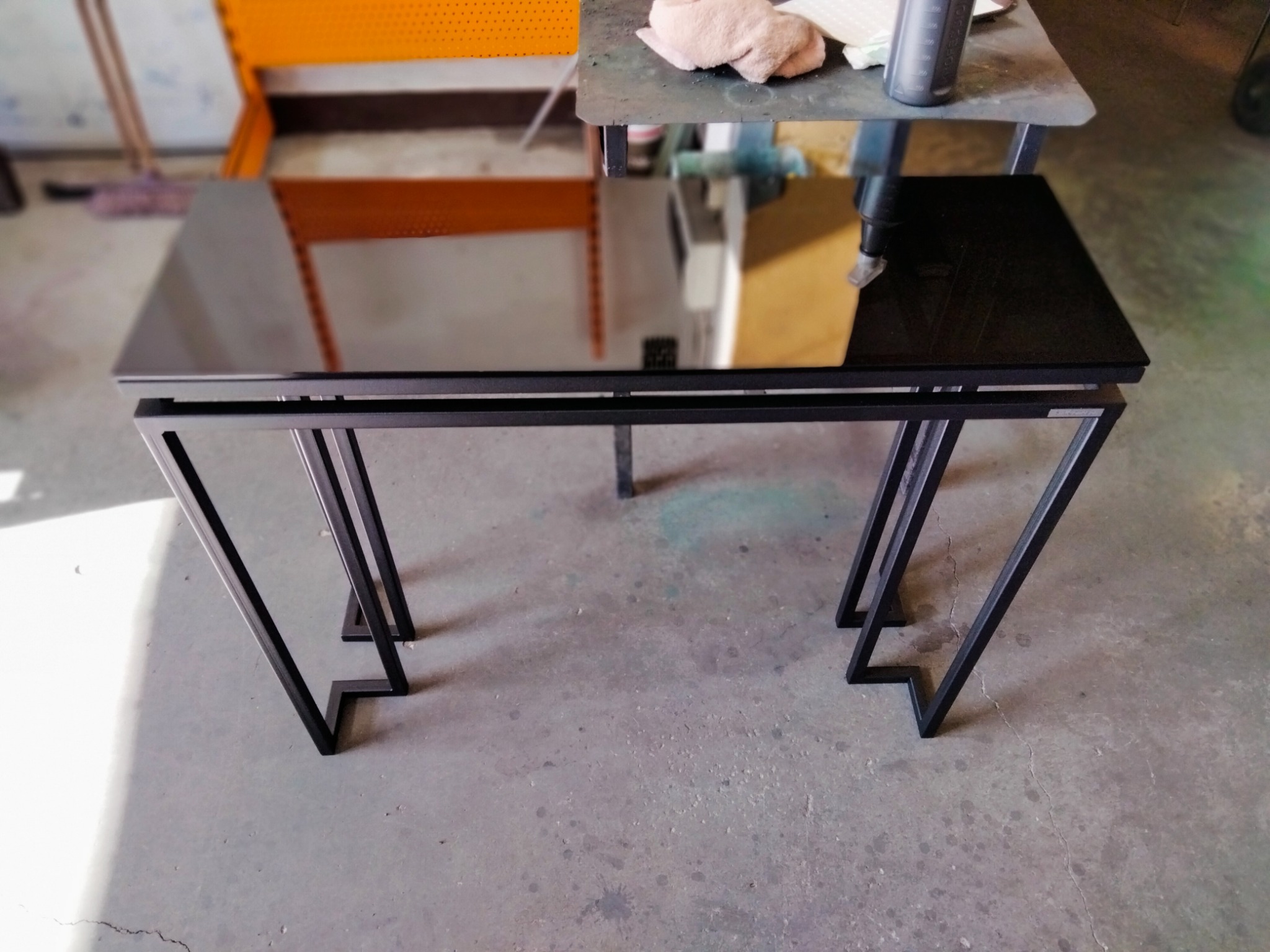 Stolik kawowy z czarnym, szklanym blatem i spawanej, geometrycznej konstrukcji nóg z profili stalowych, widoczne ślady obróbki metalu w tle.