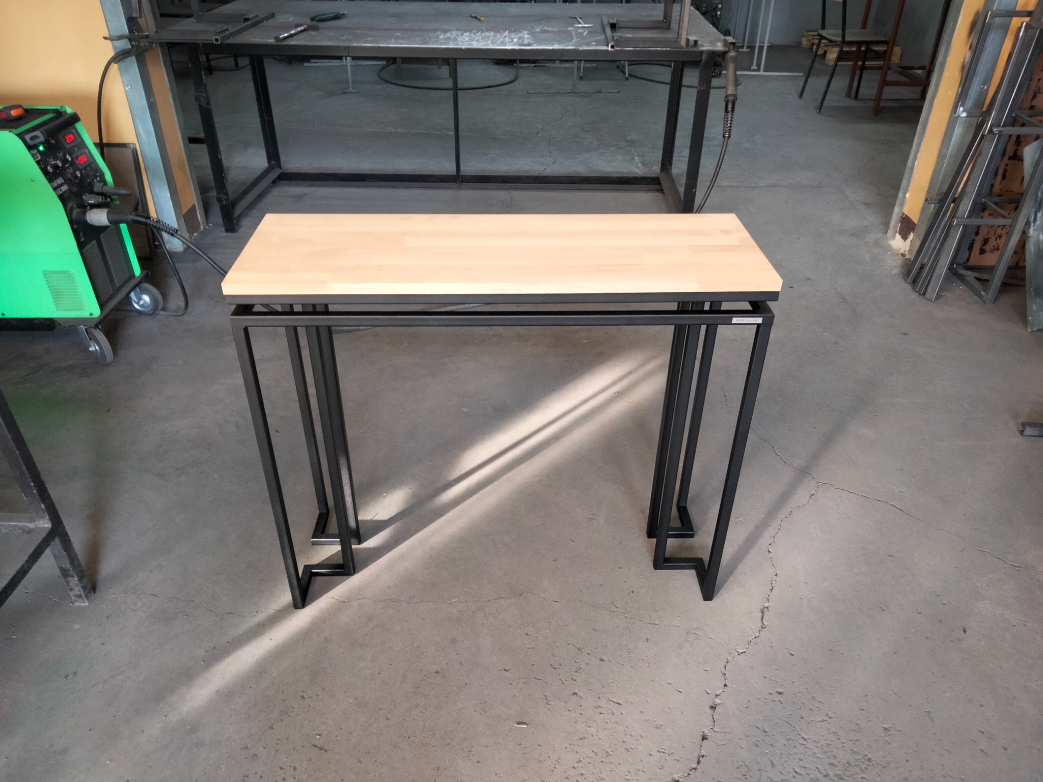 Stolik kawowy z drewnianym blatem i metalową ramą, wykonany w warsztacie, z widocznym spawarką w tle.