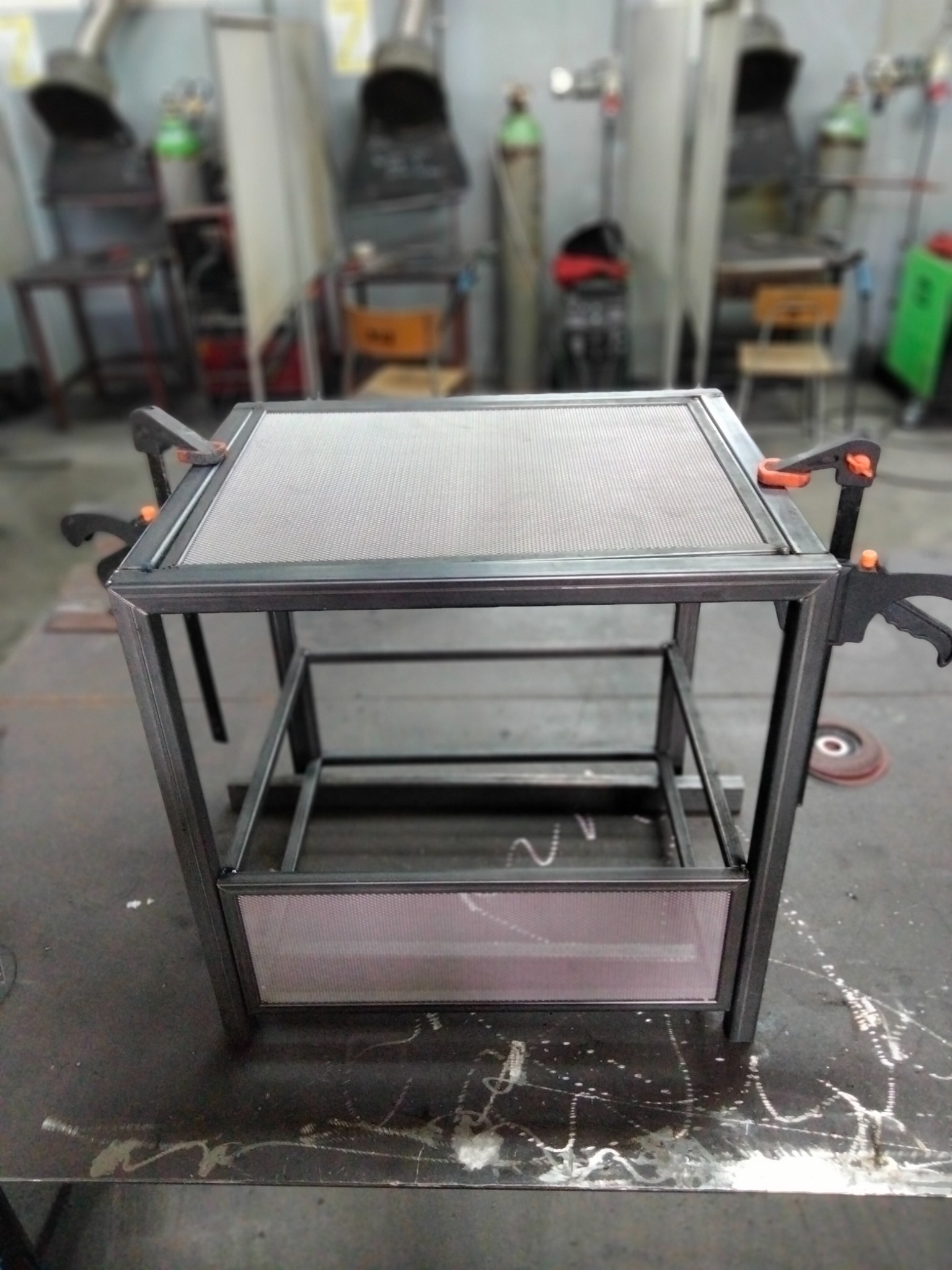 Metalowa konstrukcja stolika z siatką, w trakcie montażu, zabezpieczona klamrami stolarskimi, na tle warsztatu.