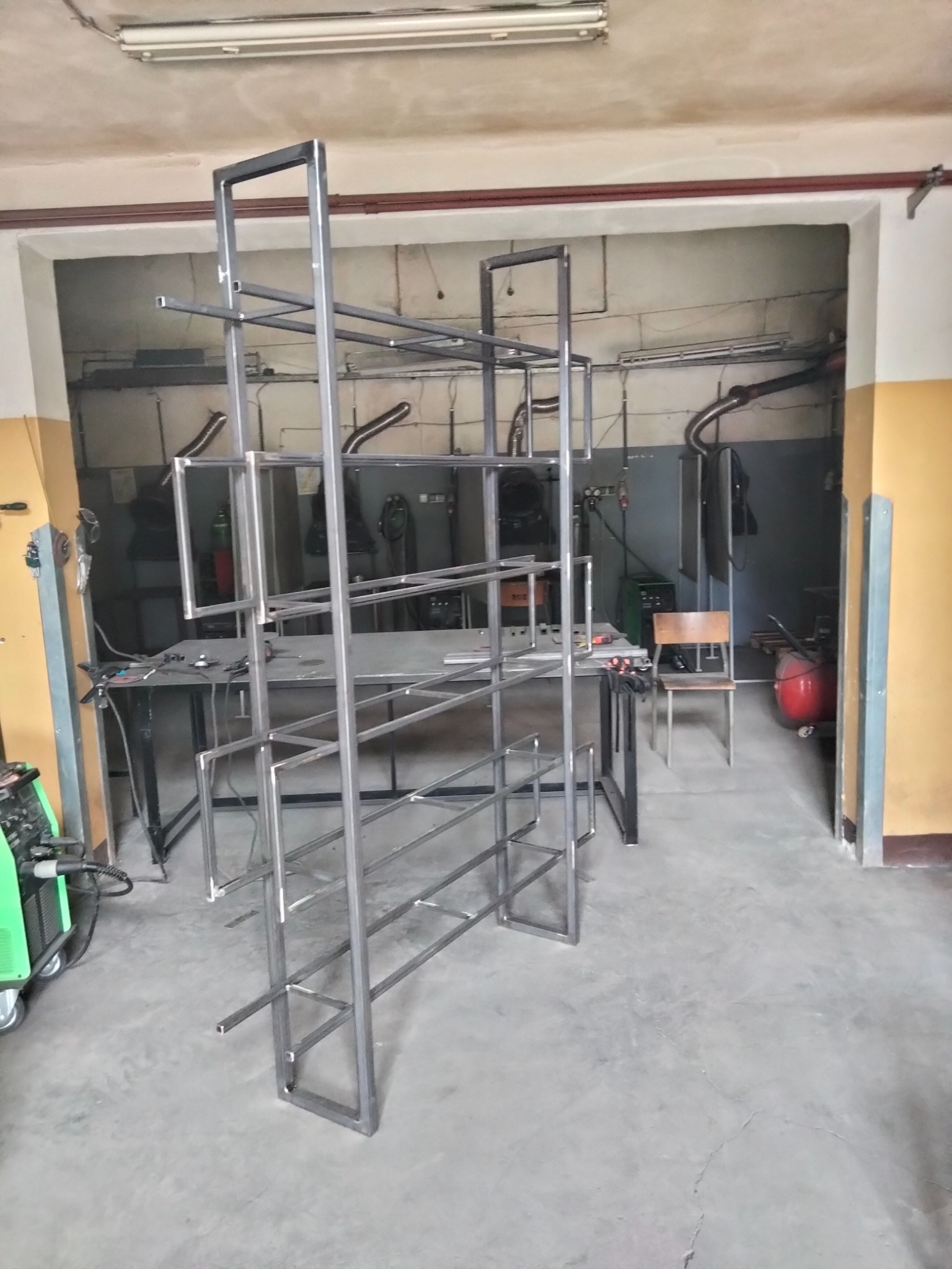 Metalowa konstrukcja regału w trakcie budowy w warsztacie, widoczny sprzęt spawalniczy w tle.