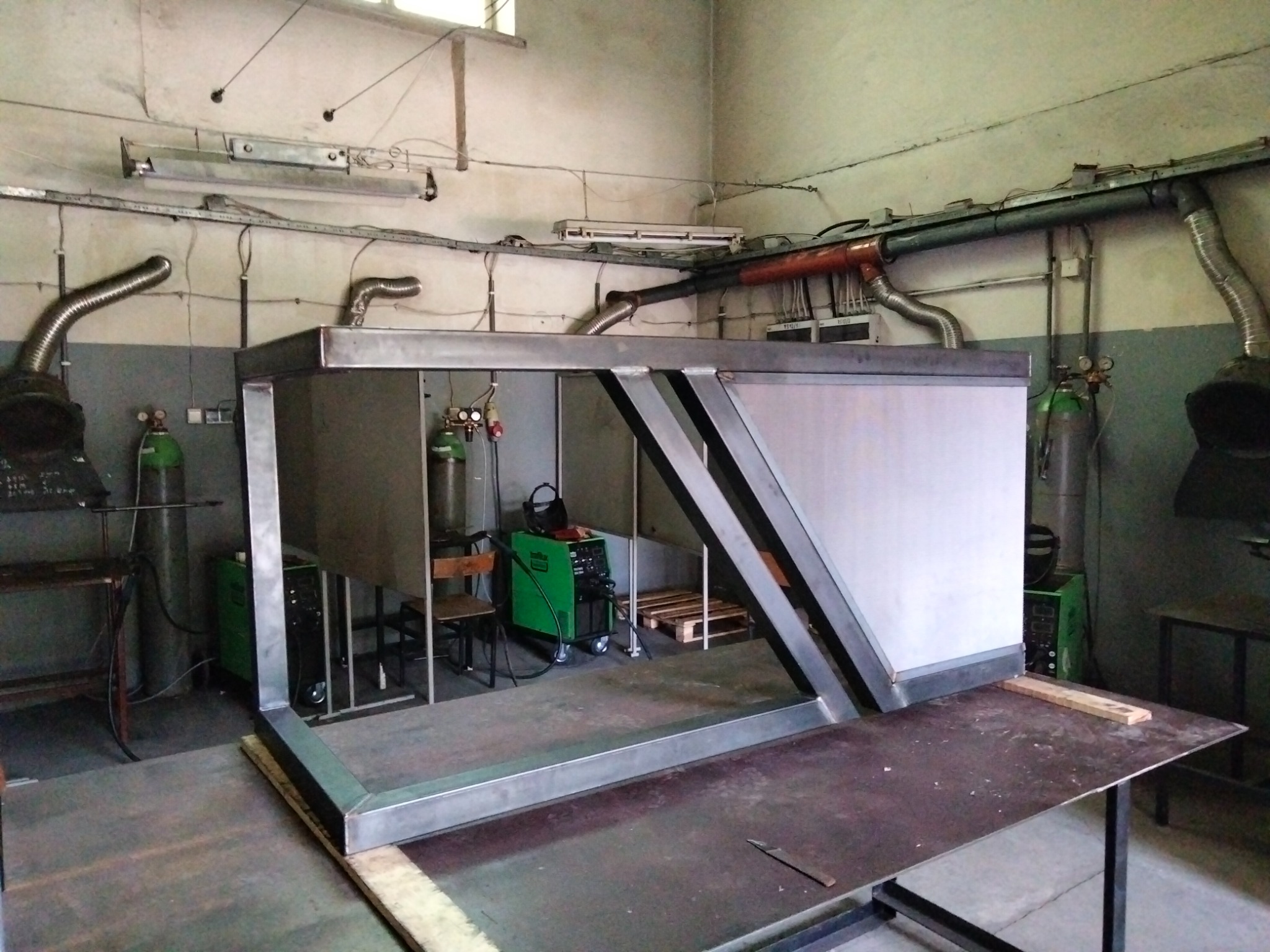 Metalowa konstrukcja z profili spawanych, częściowo osłonięta blachą, w warsztacie z instalacją wentylacyjną i butlami gazowymi.