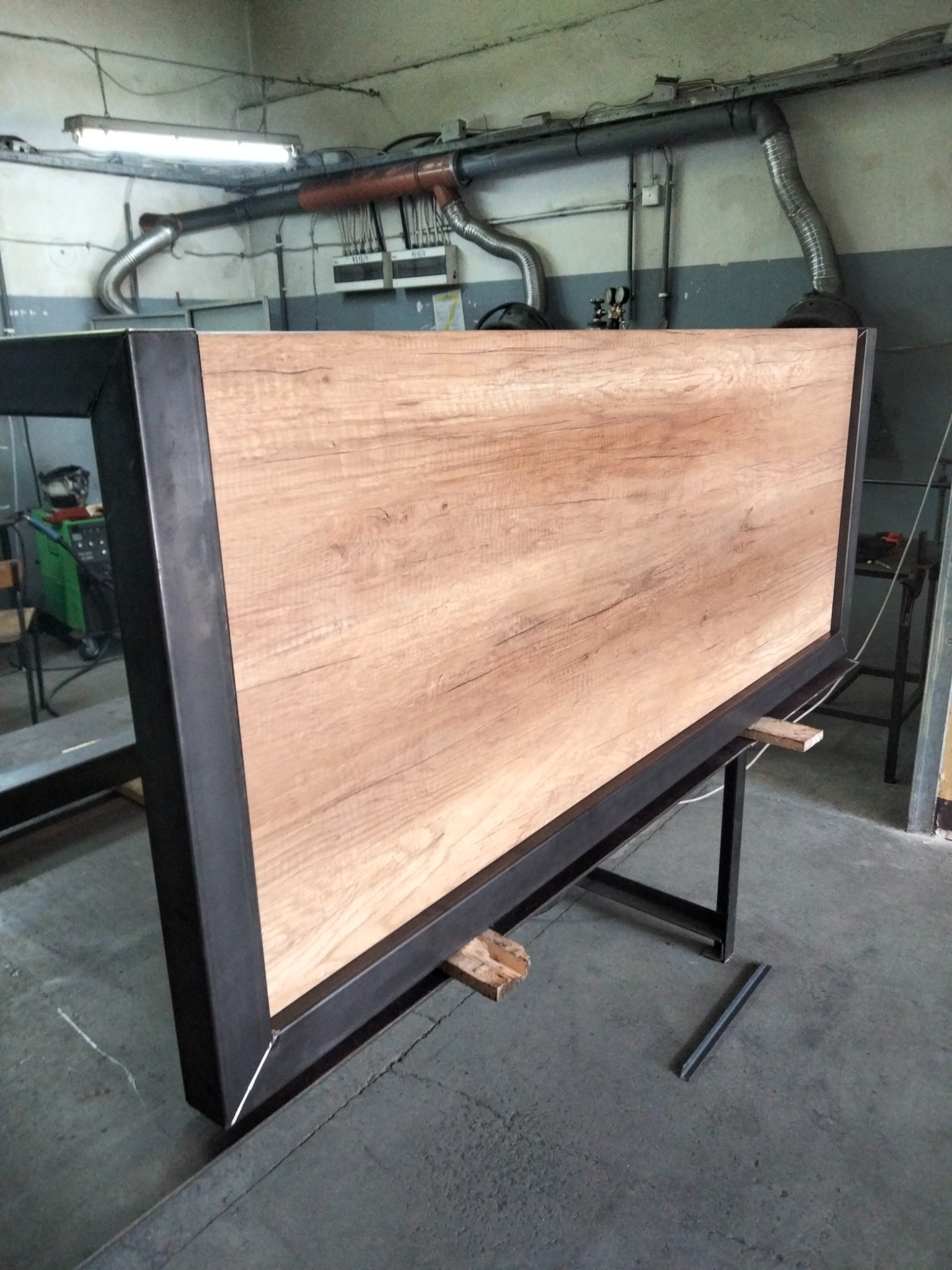 Rama z czarnego metalu otaczająca płytę drewnopodobną, wsparta na metalowym stelażu w warsztacie.