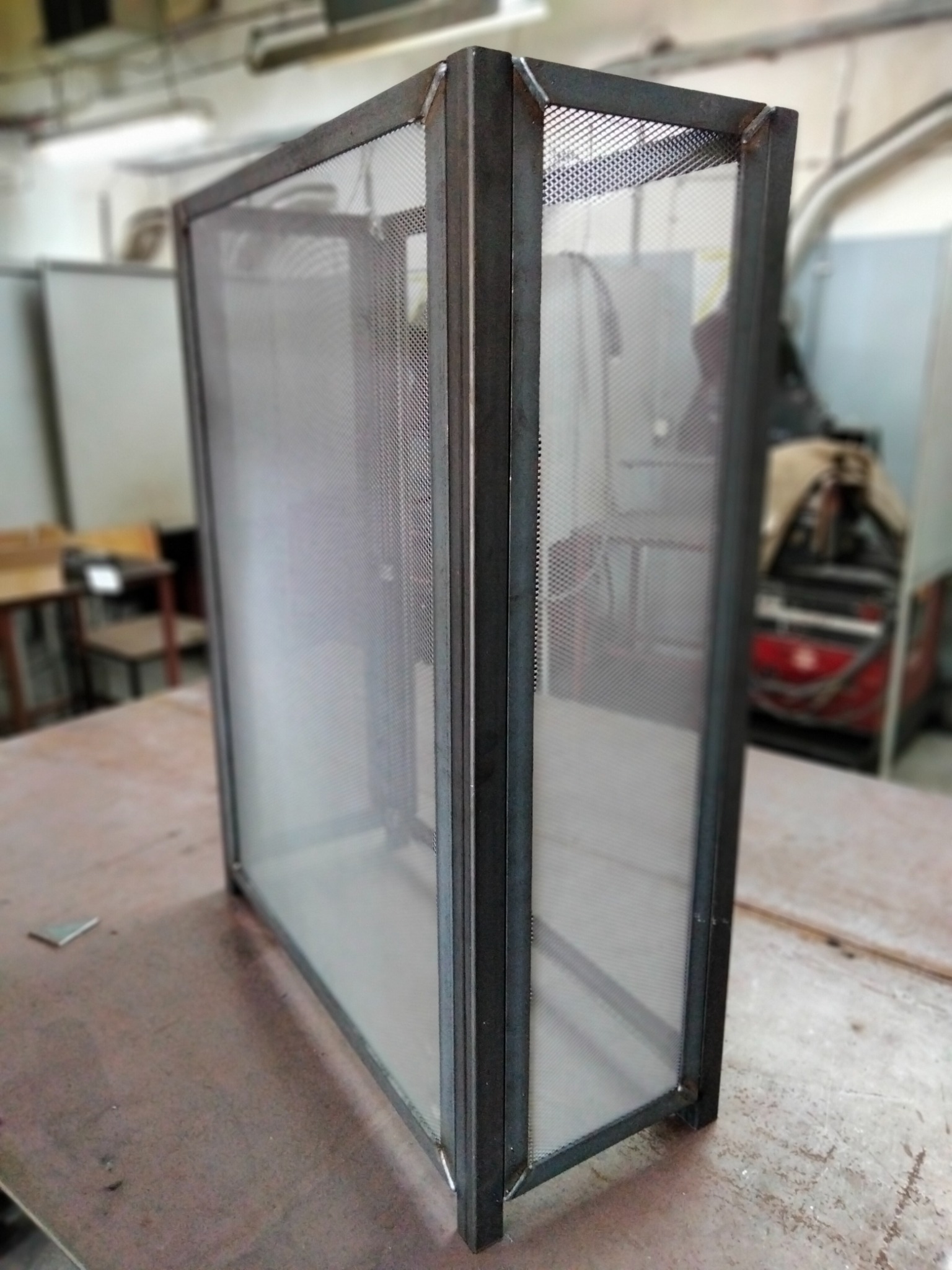 Spawana metalowa konstrukcja z siatką, widoczne spawy narożne, w tle warsztat z maszyną spawalniczą.