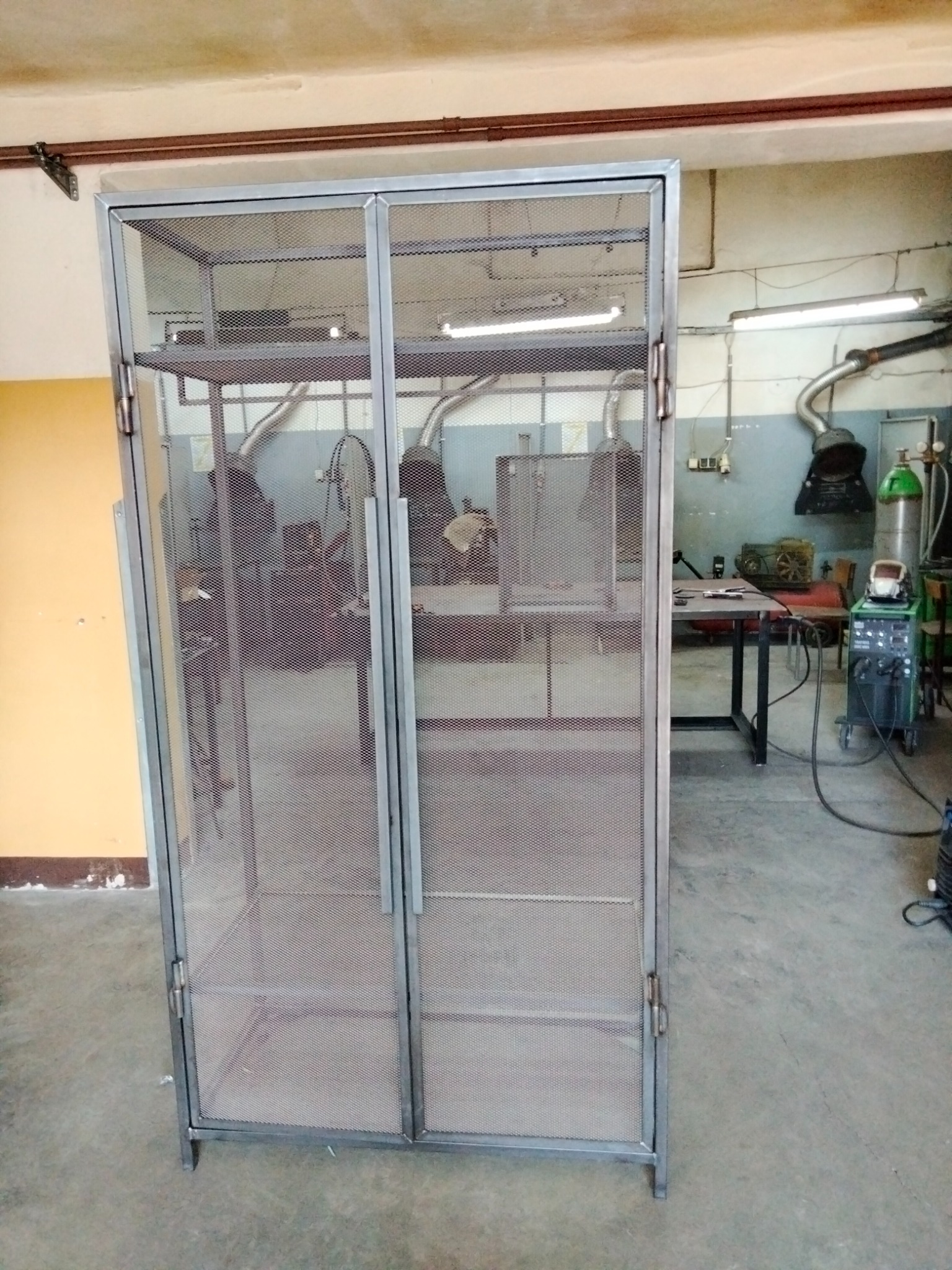 Metalowa szafa z siatki, widoczne spawane elementy konstrukcyjne, w tle warsztat ze sprzętem spawalniczym.