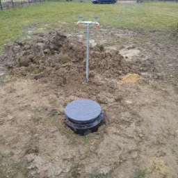 Instalacja wodna z szarą rurą i trzema kranami na tle ziemi i trawy, widoczny czarny zbiornik z szarą pokrywą, w tle ogrodzenie i samochód.