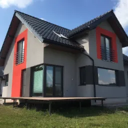 Nowoczesny dom jednorodzinny z szarą elewacją i czerwonymi akcentami wokół okien, czarną dachówką i dużymi oknami, widoczny taras z desek.