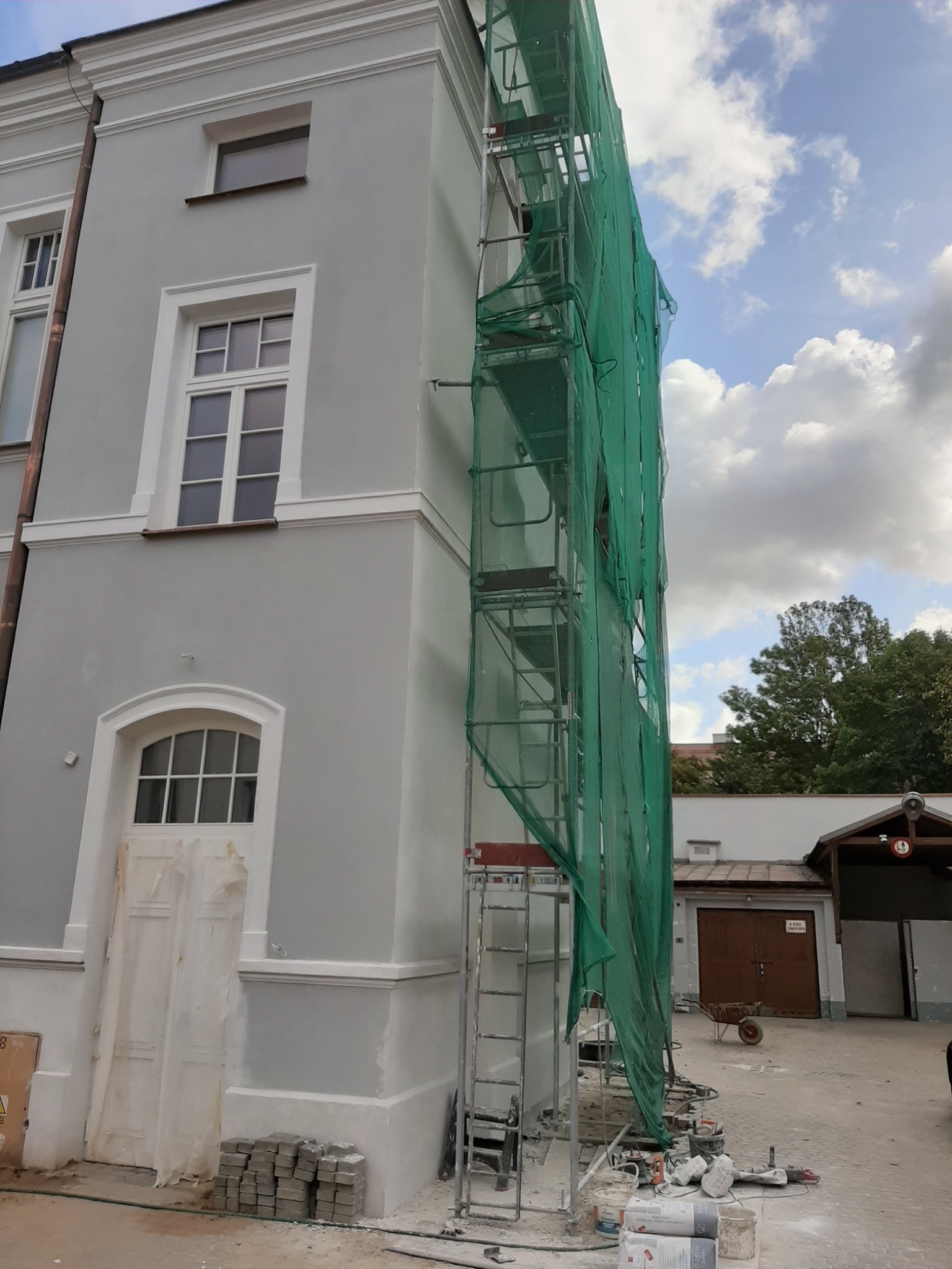 Szara elewacja budynku w trakcie tynkowania, osłonięta zieloną siatką na rusztowaniu, widoczne narzędzia i materiały budowlane, kostka brukowa przy wejściu.