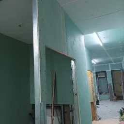 Korytarz w trakcie remontu z widocznymi płytami gipsowo-kartonowymi na ścianach i suficie, wyznaczonym poziomem laserowym oraz metalową konstrukcją ścianki działowej.