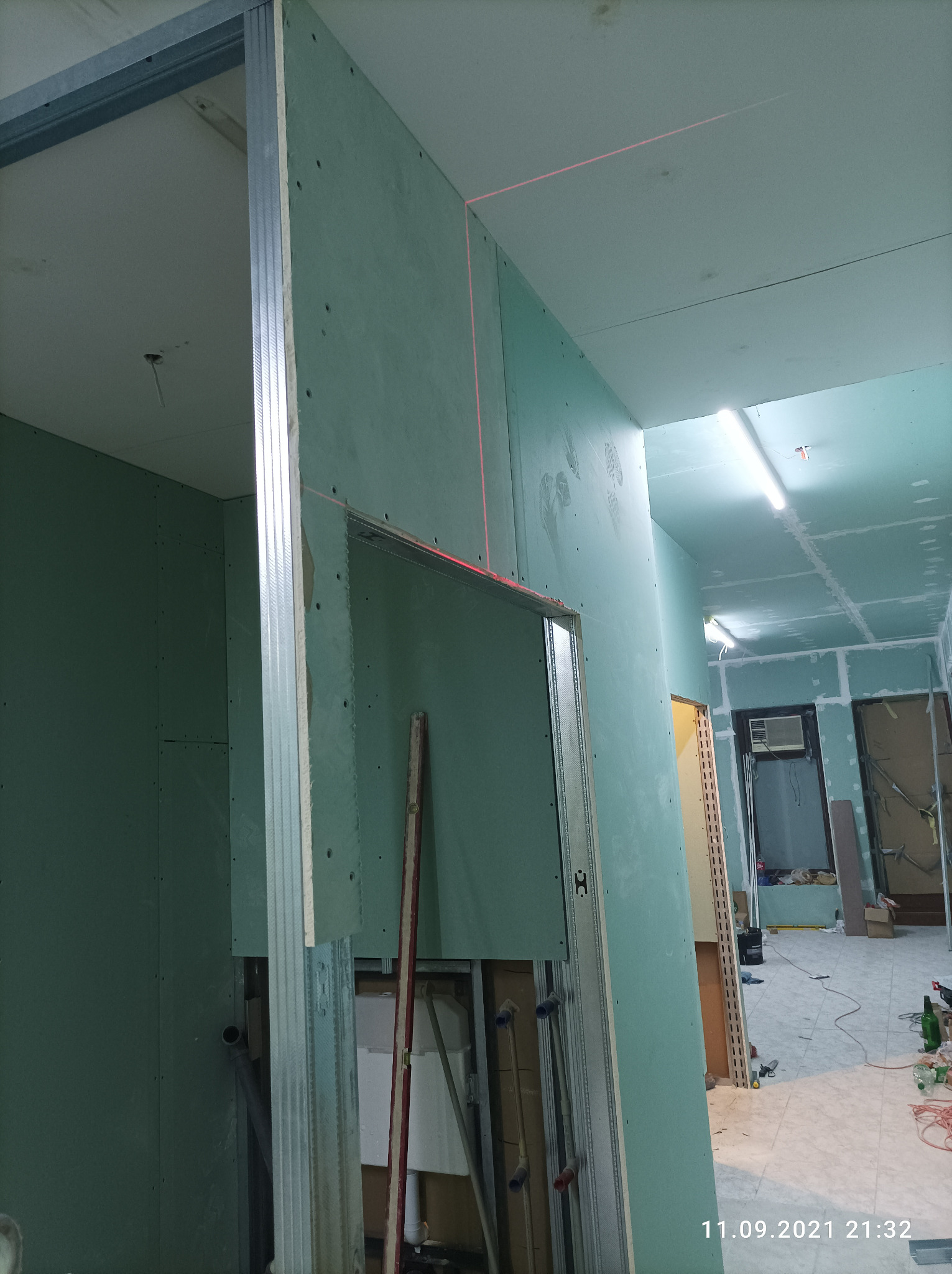 Korytarz w trakcie remontu z widocznymi płytami gipsowo-kartonowymi na ścianach i suficie, wyznaczonym poziomem laserowym oraz metalową konstrukcją ścianki działowej.