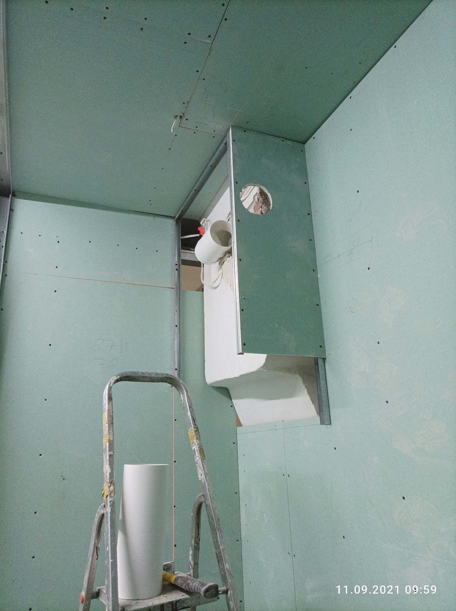 Wnętrze w trakcie prac wykończeniowych: ściany z płyt kartonowo-gipsowych w odcieniach zieleni, aluminiowa drabina z młotkiem na stopniu, widoczny fragment instalacji wentylacyjnej z otworem w płycie.