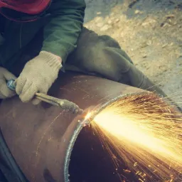 Spawacz w czerwonym kasku tnący metalową rurę za pomocą palnika acetylenowo-tlenowego, iskry rozbryzgują się na boki.