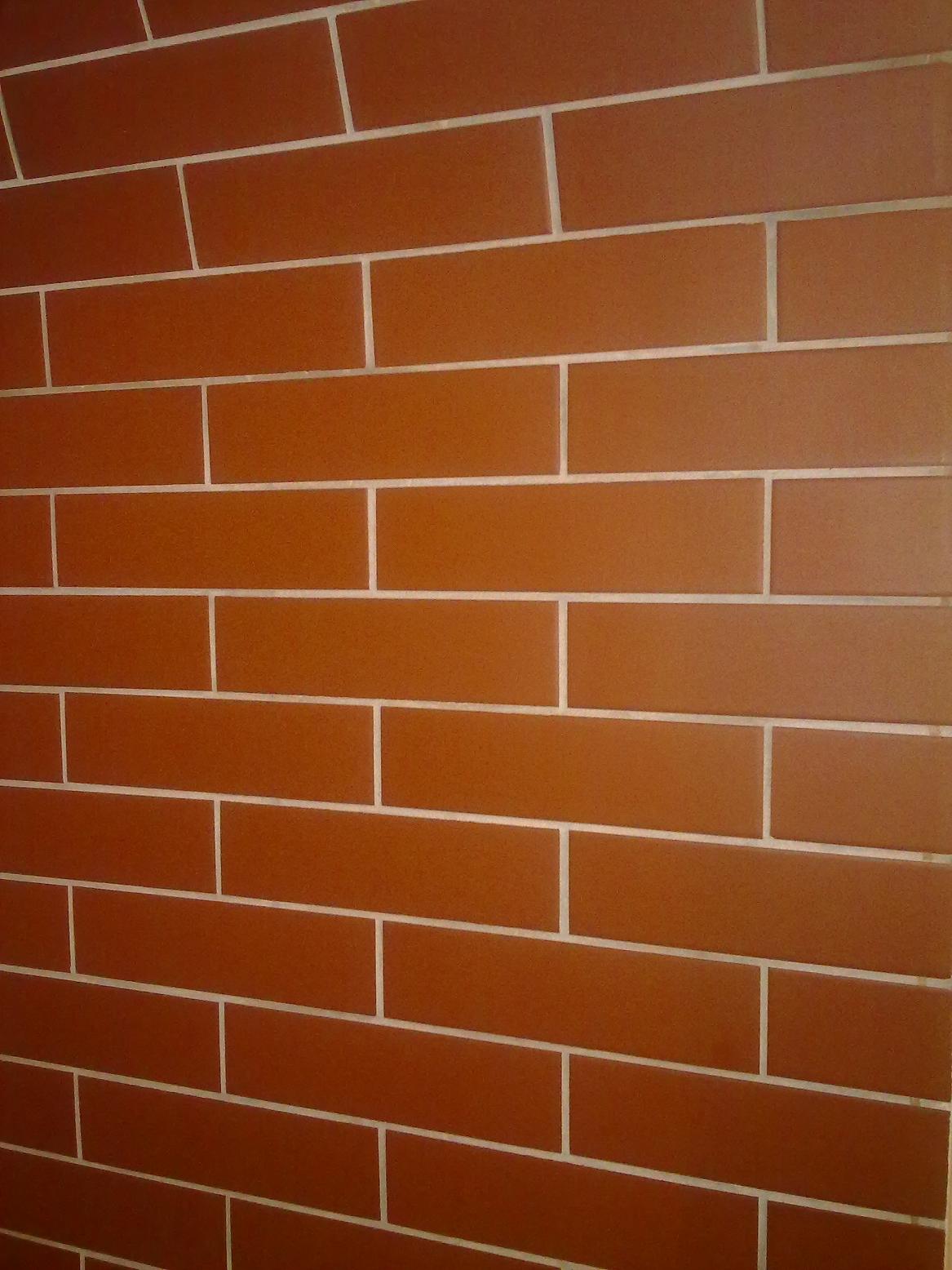 Ściana wyłożona brązowymi, prostokątnymi płytkami z jasną fugą, ułożonymi w regularny wzór.