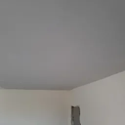 Pomalowany na szaro sufit w trakcie remontu mieszkania, widoczne ślady po wałku malarskim i niedomalowane fragmenty przy ścianie, zarys otworu w ścianie.