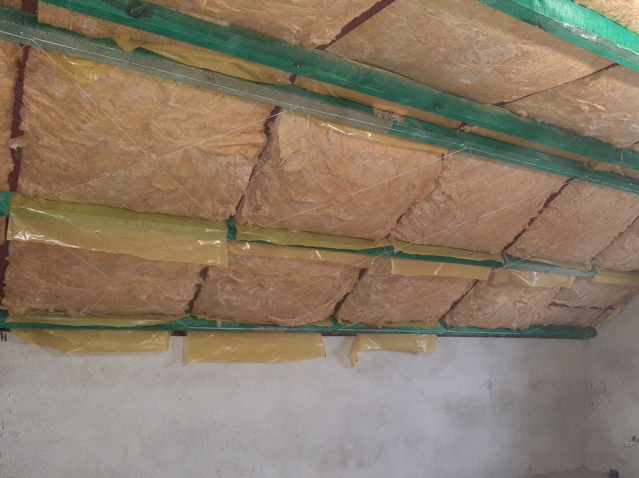 Izolacja termiczna poddasza z wełny mineralnej, zabezpieczona folią, widoczne zielone belki konstrukcyjne i sznurki mocujące.