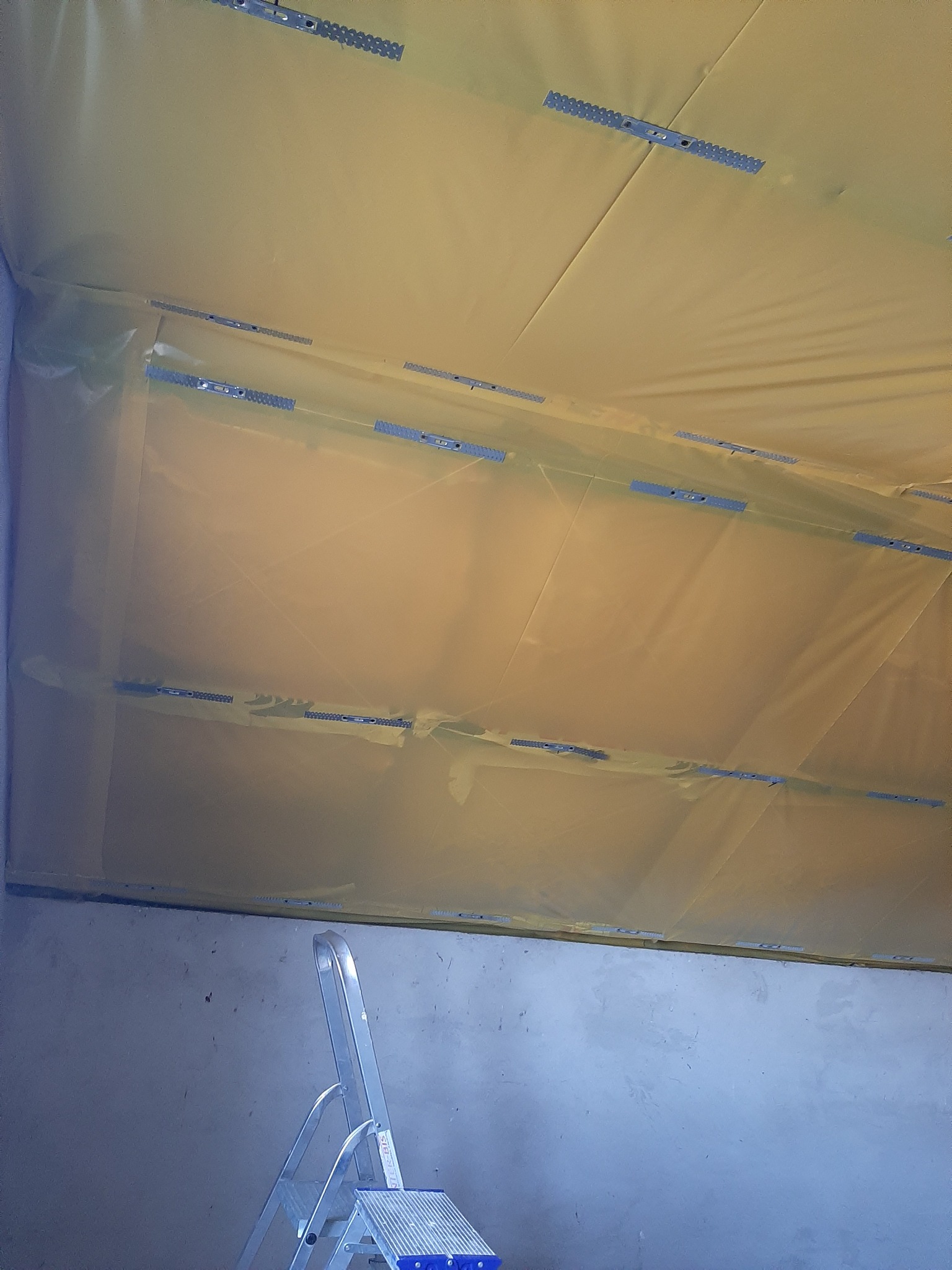 Żółta folia paroizolacyjna zamocowana do sufitu za pomocą metalowych listew z ząbkami, widoczna drabina aluminiowa.