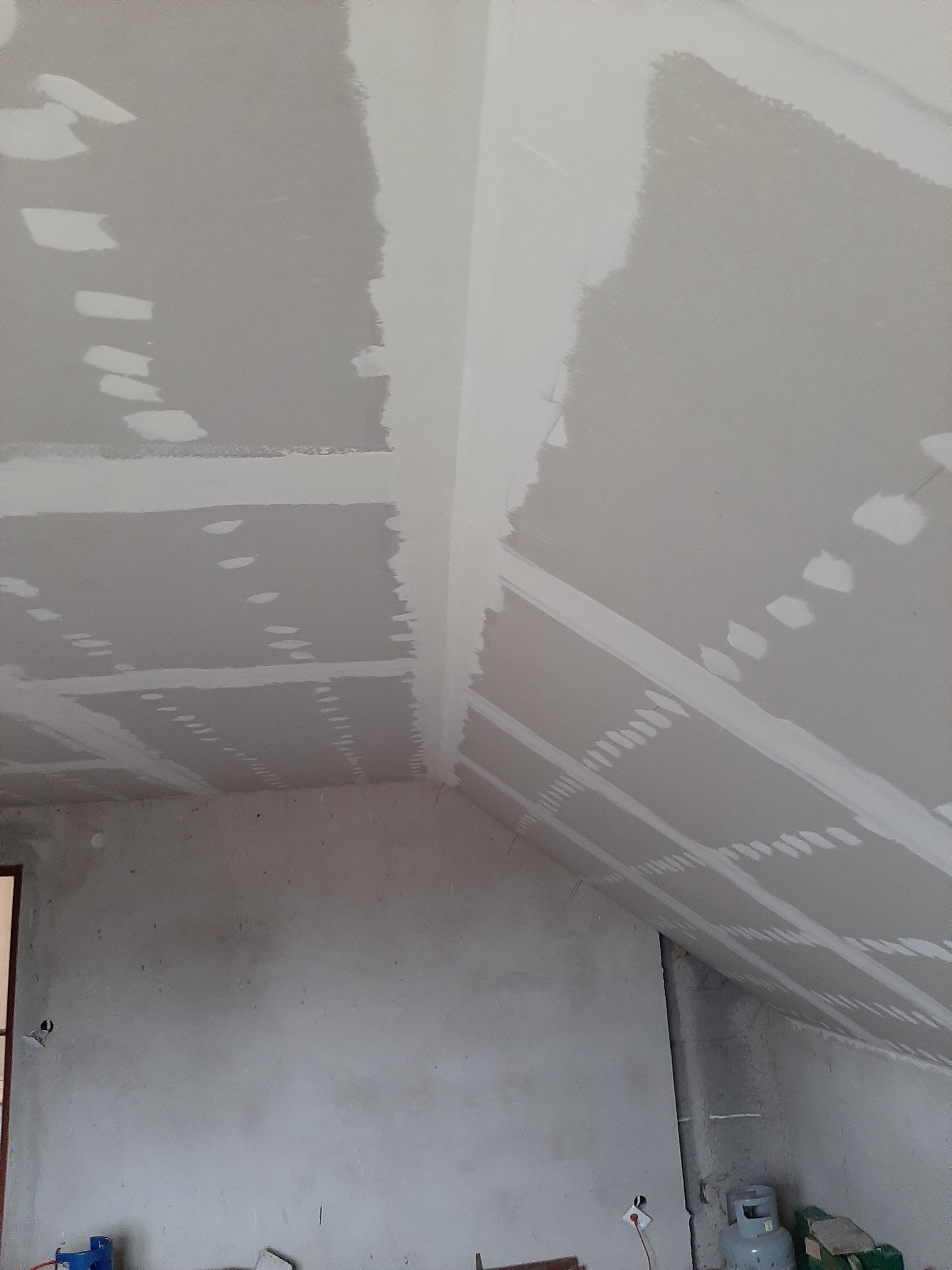 Sufit poddasza z płyt gipsowo-kartonowych w trakcie szpachlowania łączeń, widoczne białe paski wzmacniające i miejsca po wkrętach, surowe ściany w tle.