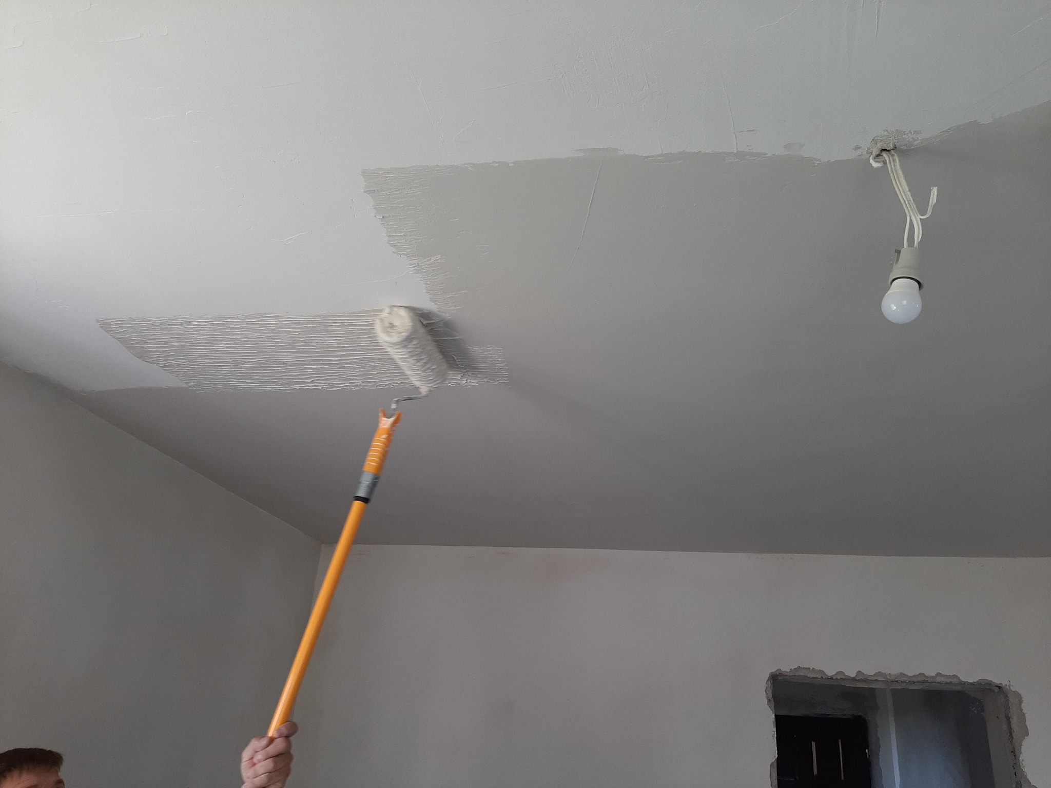 Malowanie sufitu wałkiem z teleskopową rączką w szarym kolorze, widoczna niedokończona powierzchnia i wisząca żarówka bez klosza.