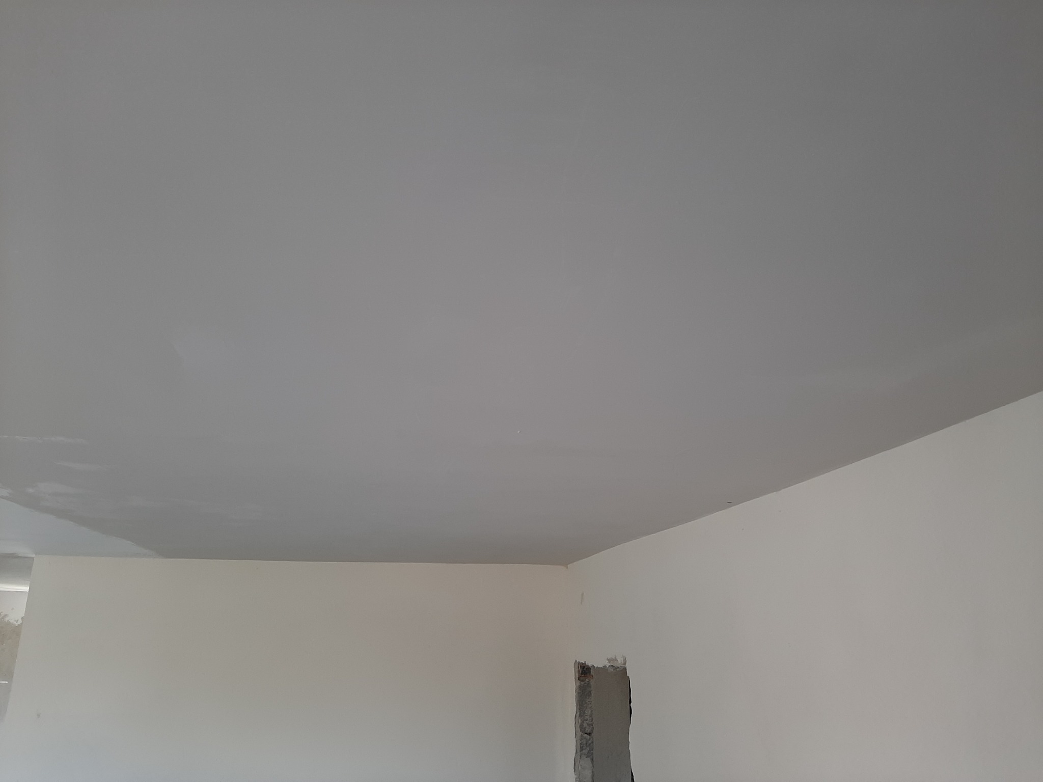 Pomalowany na szaro sufit w trakcie remontu mieszkania, widoczne ślady po wałku malarskim i niedomalowane fragmenty przy ścianie, zarys otworu w ścianie.