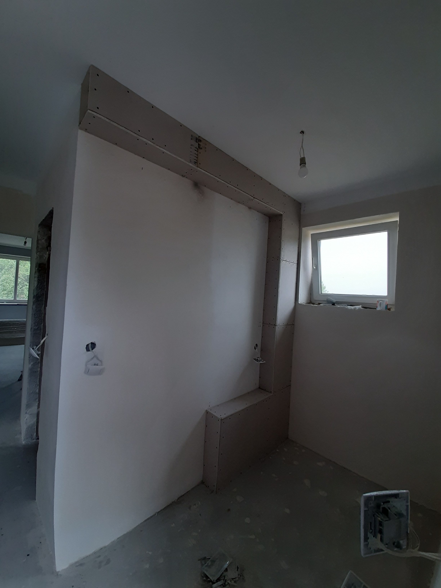 Wnętrze w trakcie remontu: ściana z płyt gipsowo-kartonowych z widocznymi łączeniami na stelażu, surowa żarówka zwisająca z sufitu, okno z parapetem, odsłonięte gniazdko elektryczne, podłoga...