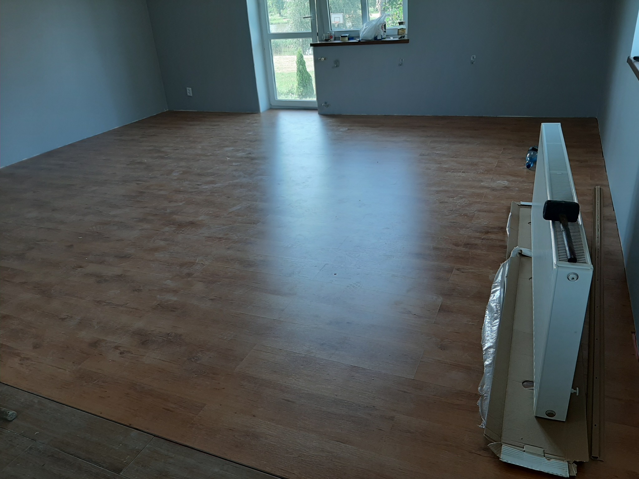 Układanie paneli podłogowych w pustym, szarym pokoju; w tle okno z widokiem na zieleń, obok ściany grzejnik w kartonie.