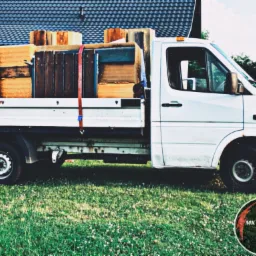 Biały samochód dostawczy z otwartą przestrzenią ładunkową wypełnioną meblami i zabezpieczony czerwonymi pasami, zaparkowany na trawniku przed budynkiem z ciemnym dachem, z widocznym logo firmy...