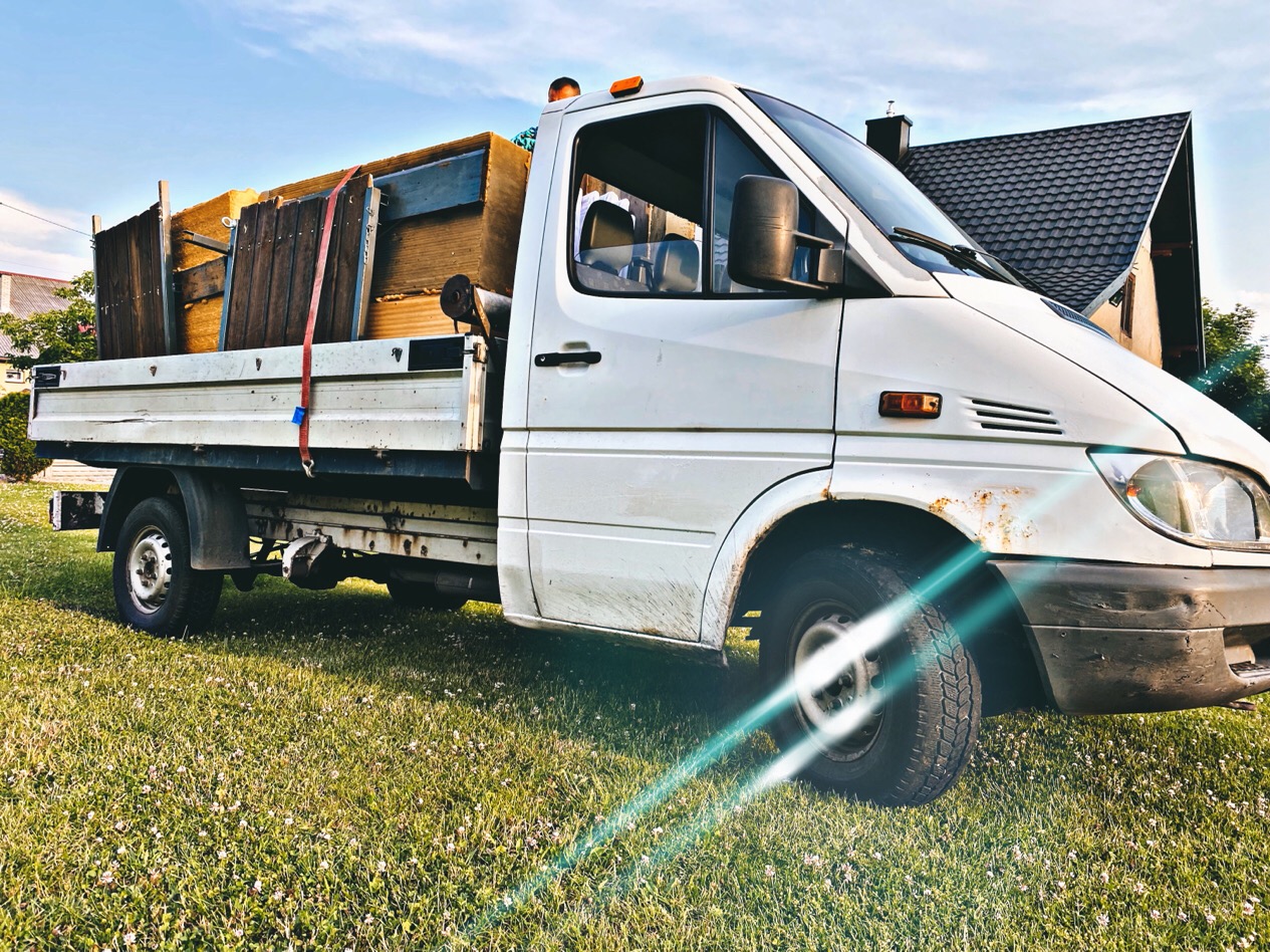 Biały samochód dostawczy z otwartą skrzynią ładunkową, załadowany drewnianymi elementami i płytami, zabezpieczony czerwonym pasem, stojący na trawniku przed domem.