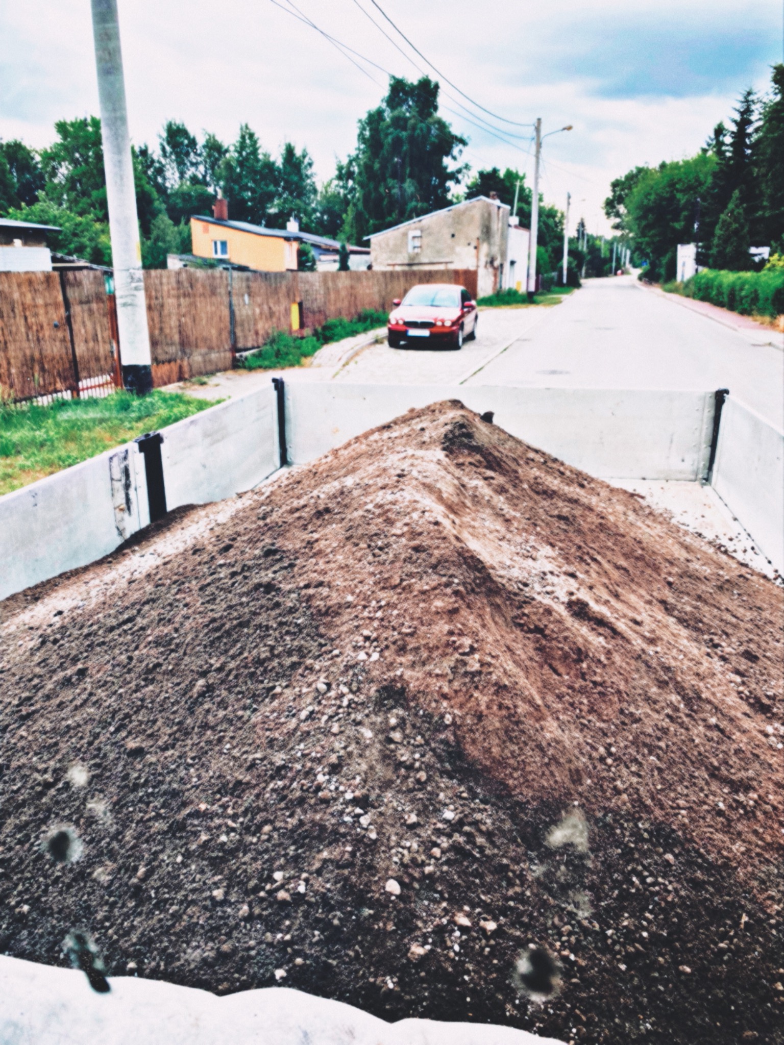 Kupa ziemi w skrzyni ładunkowej samochodu ciężarowego, w tle ulica z czerwonym samochodem osobowym i zabudowaniami.