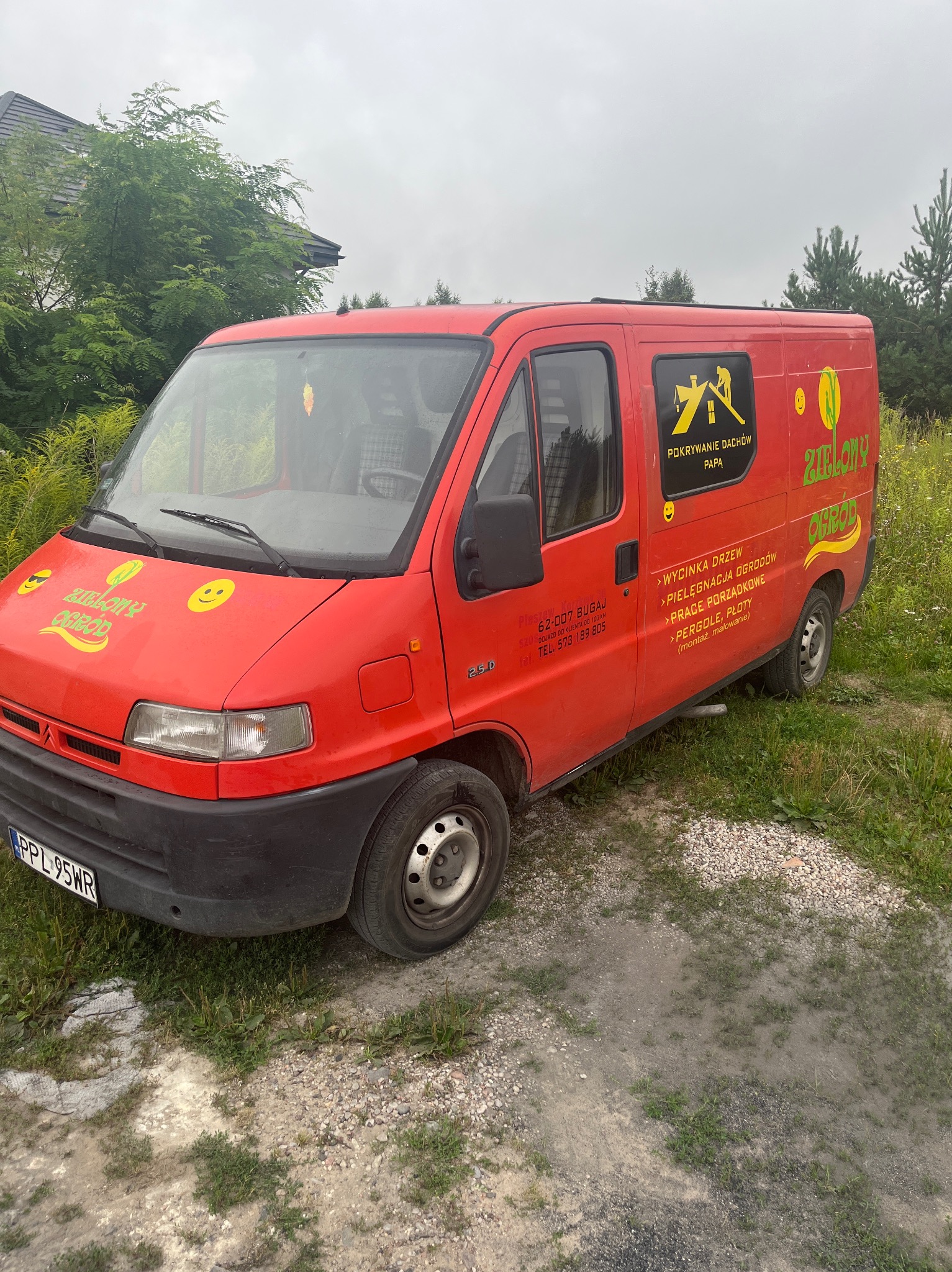 Czerwony samochód dostawczy z reklamą firmy świadczącej usługi ogrodnicze i dekarskie, zaparkowany na żwirowym podłożu w pobliżu zieleni.