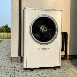Zewnętrzna jednostka urządzenia grzewczego marki Bosch zamontowana przy ścianie budynku z widocznymi rurami przyłączeniowymi i niewielkim zabrudzeniem na obudowie.