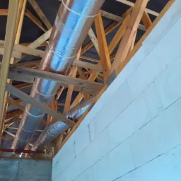 Dwa aluminiowe przewody wentylacyjne zamontowane pod drewnianą konstrukcją dachu w budynku z białych bloczków.