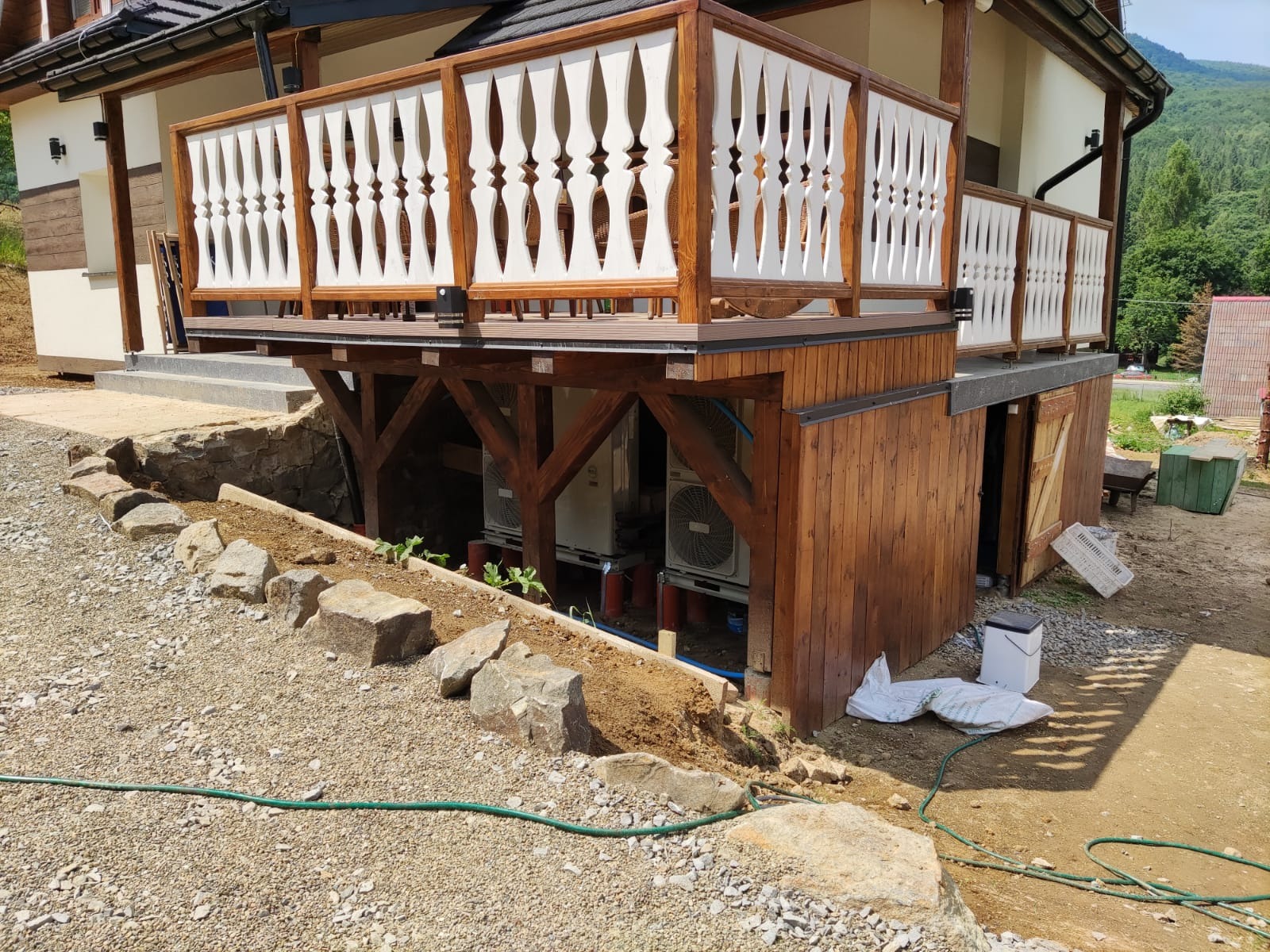 Zewnętrzne jednostki klimatyzacyjne pod drewnianym tarasem z ozdobną balustradą, widoczne rury i instalacje, otoczenie w trakcie zagospodarowywania, kamienie i ziemia.