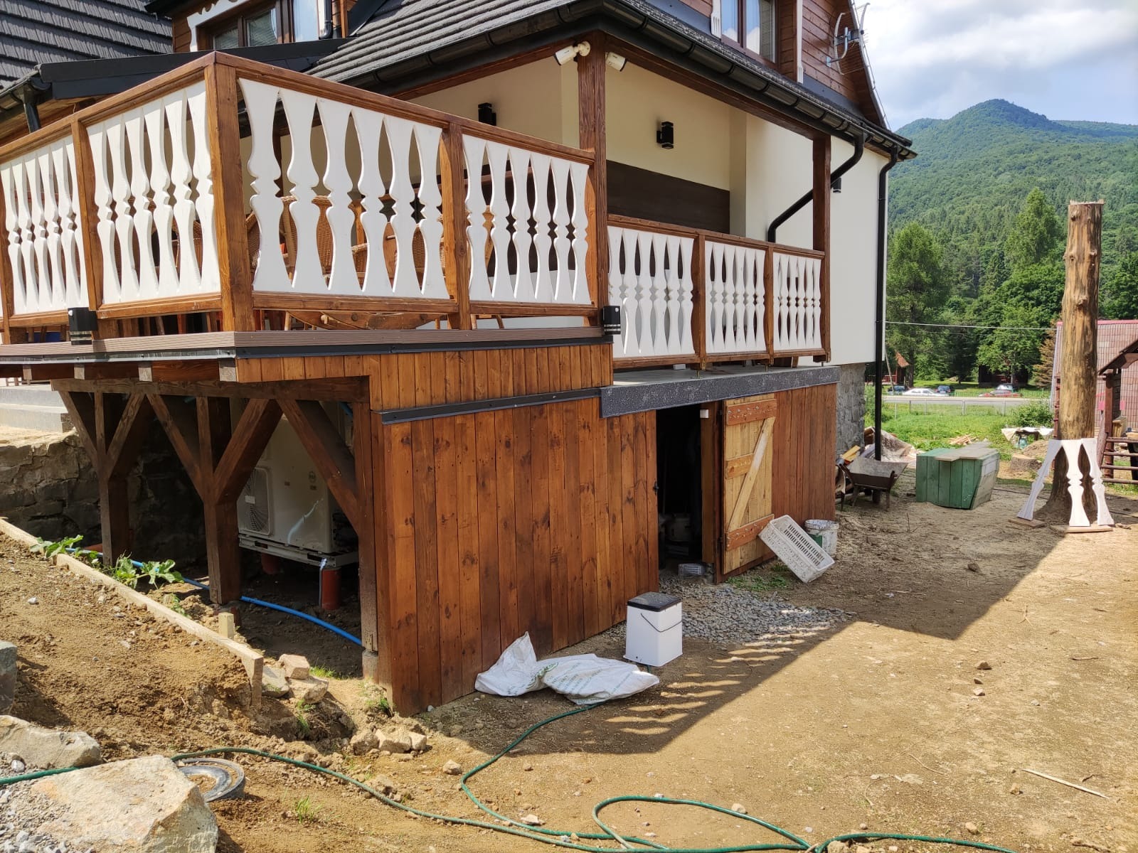 Dom z drewnianą elewacją i balkonem z białą balustradą, w trakcie prac wykończeniowych na zewnątrz, widoczna instalacja pod spodem i materiały budowlane.