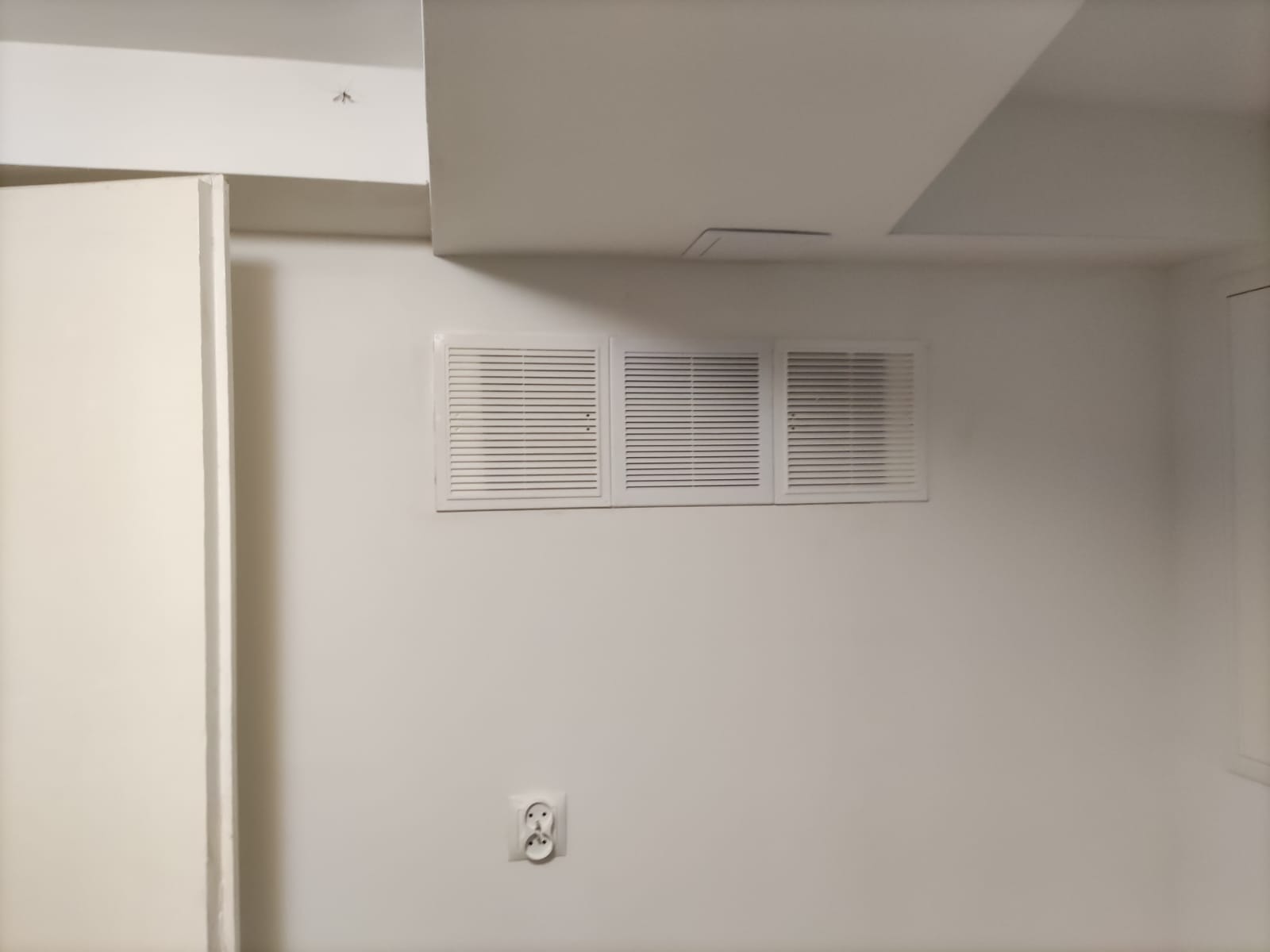 Trzy białe kratki wentylacyjne zamontowane na białej ścianie, poniżej gniazdko elektryczne.