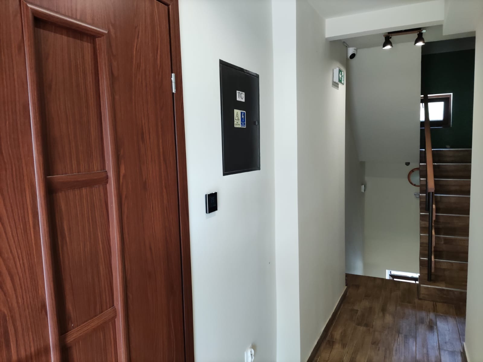 Wnętrze budynku z brązowymi drzwiami, tablicą rozdzielczą z symbolami ostrzegawczymi na białej ścianie oraz widocznymi schodami w głębi.