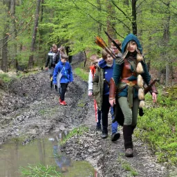 Grupa dzieci i młodzieży wędruje leśną ścieżką, na czele dziewczyna przebrana za postać z fantasy, z łukiem i strzałami.