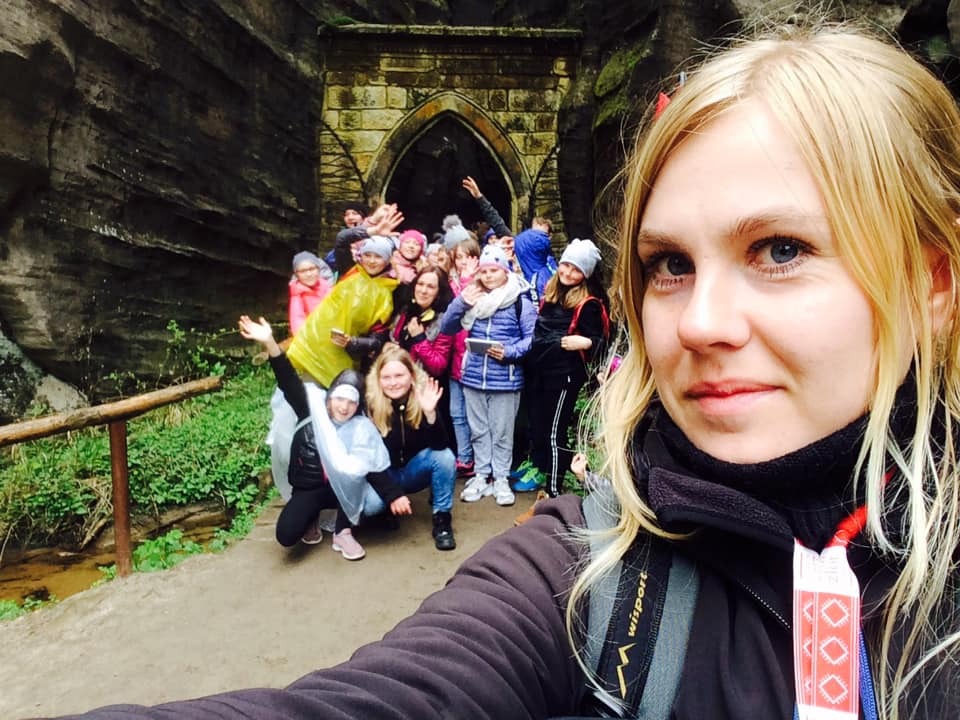 Grupa turystów pozuje do selfie na tle skalnego łuku; w pierwszym planie blondwłosa kobieta z plecakiem.